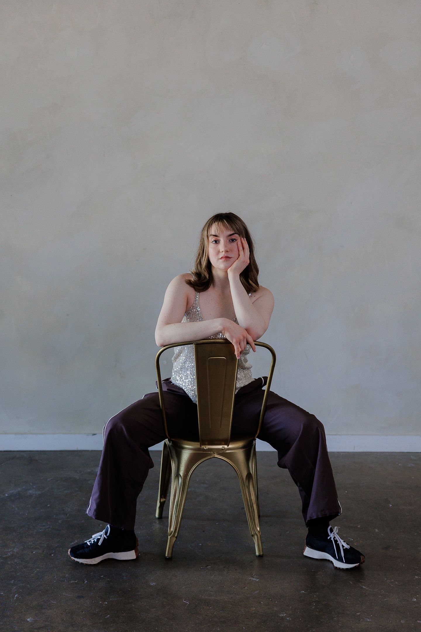 A person in dark clothing poses artistically with a metal chair against an industrial backdrop.