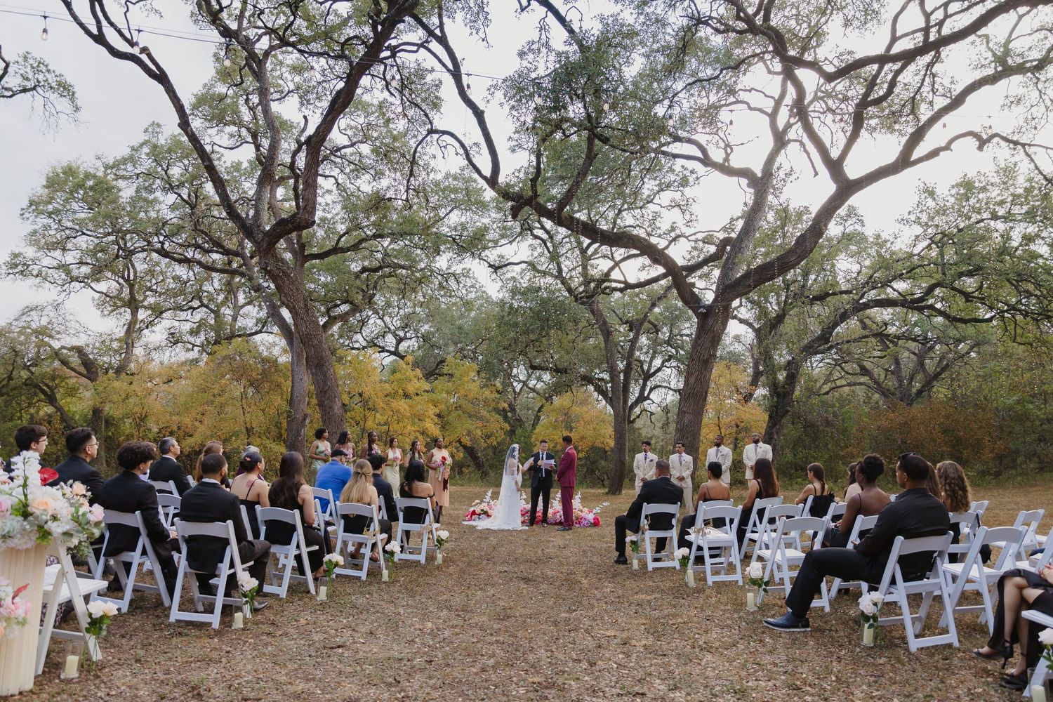 An outdoor wedding ceremony setup with white chairs arranged beneath large oak trees in a natural setting.