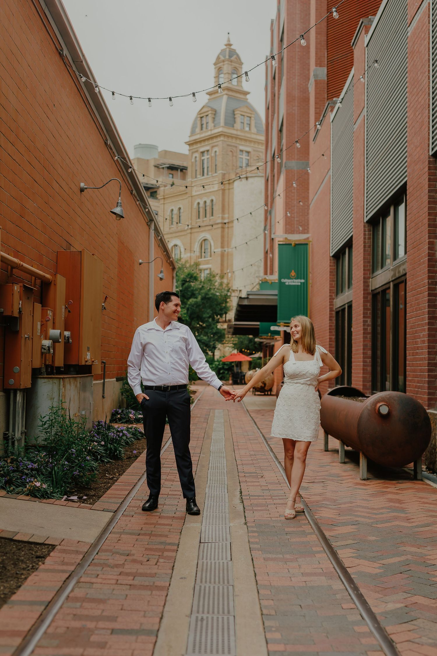A couple holds hands while walking down a brick alley with string lights and historic architecture in the background.