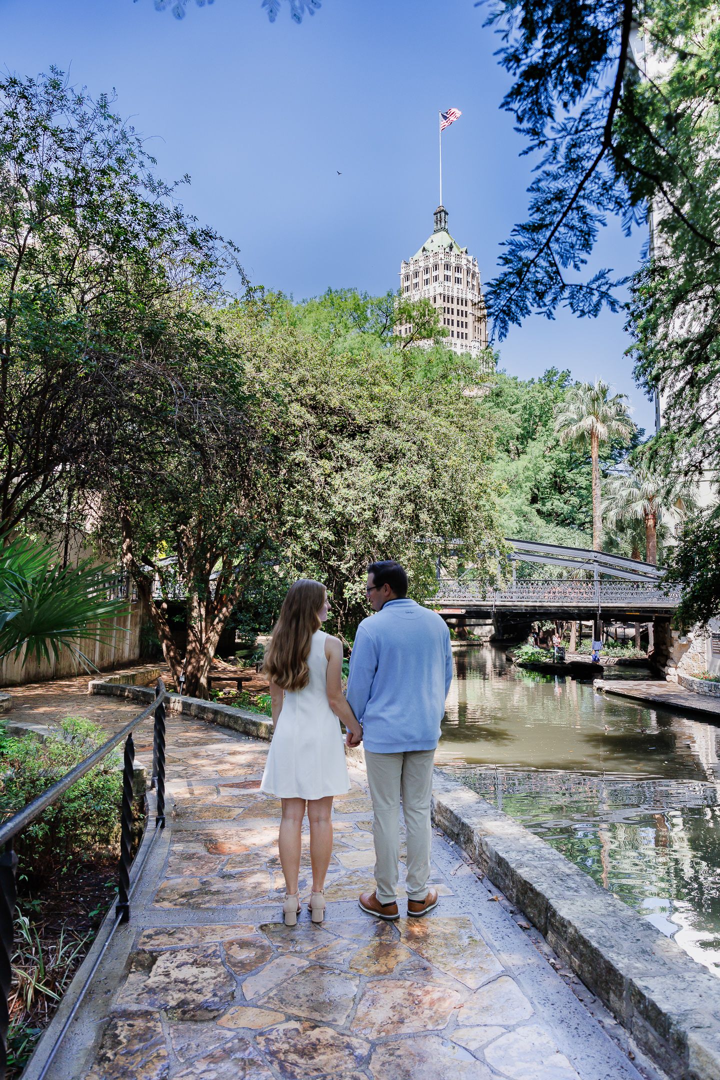 Couple holding hands strolling along the San Antonio River Walk with lush trees and Tower of the Americas in background.