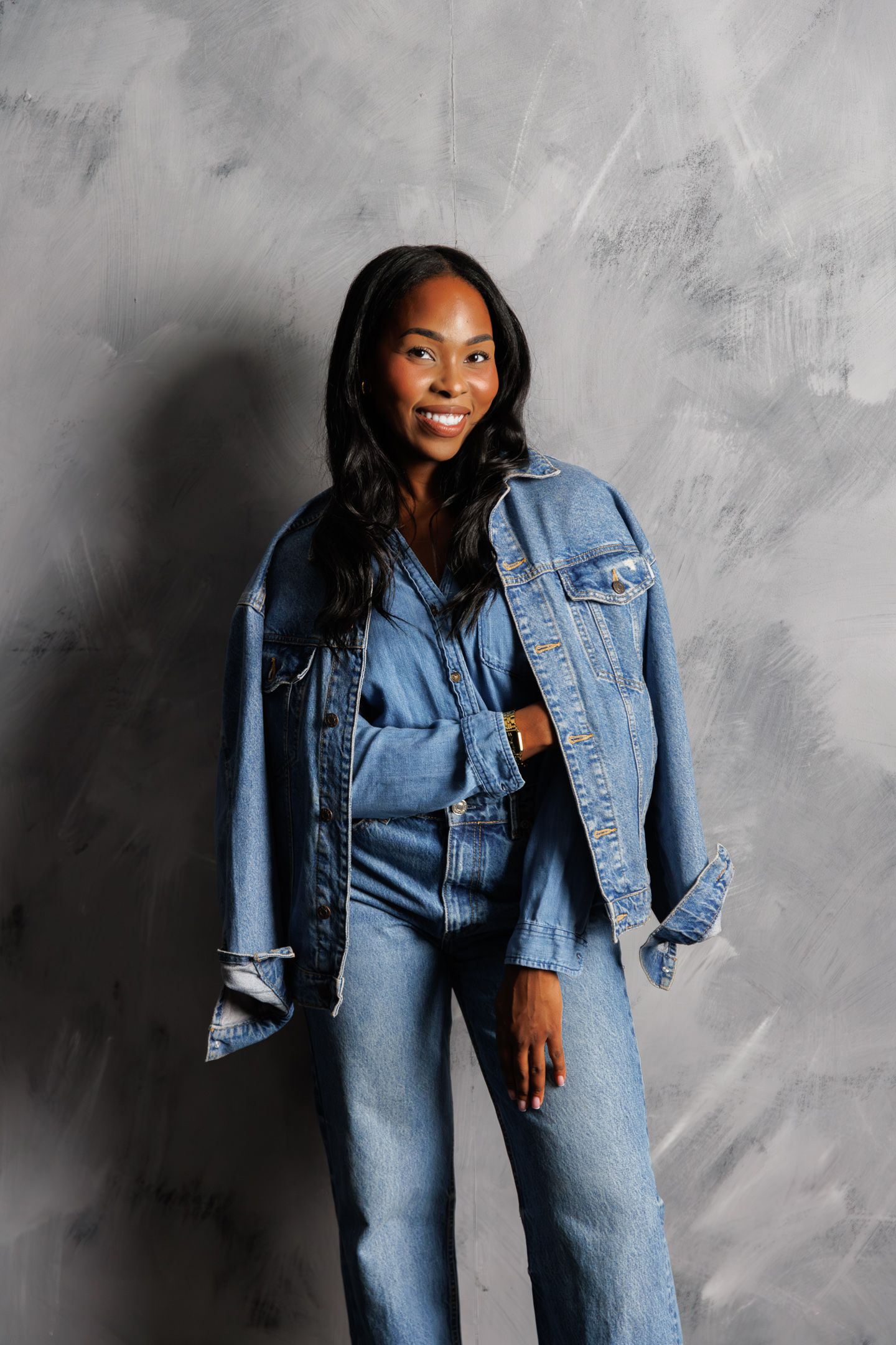 Smiling woman in a denim jacket and jeans outfit posing against a gray textured wall.