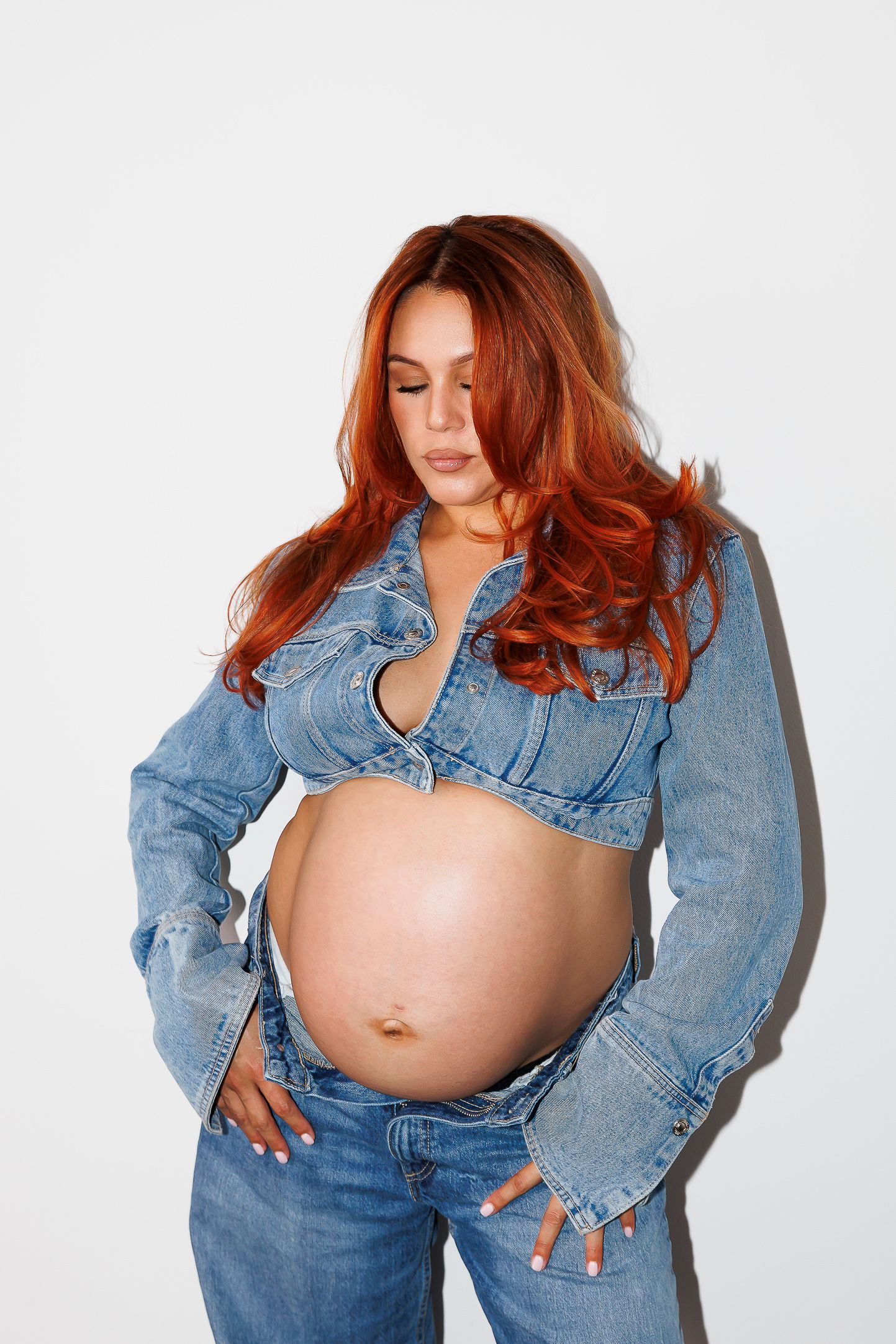 Woman in denim jacket shows bare pregnant belly against white background in artistic maternity photo series.