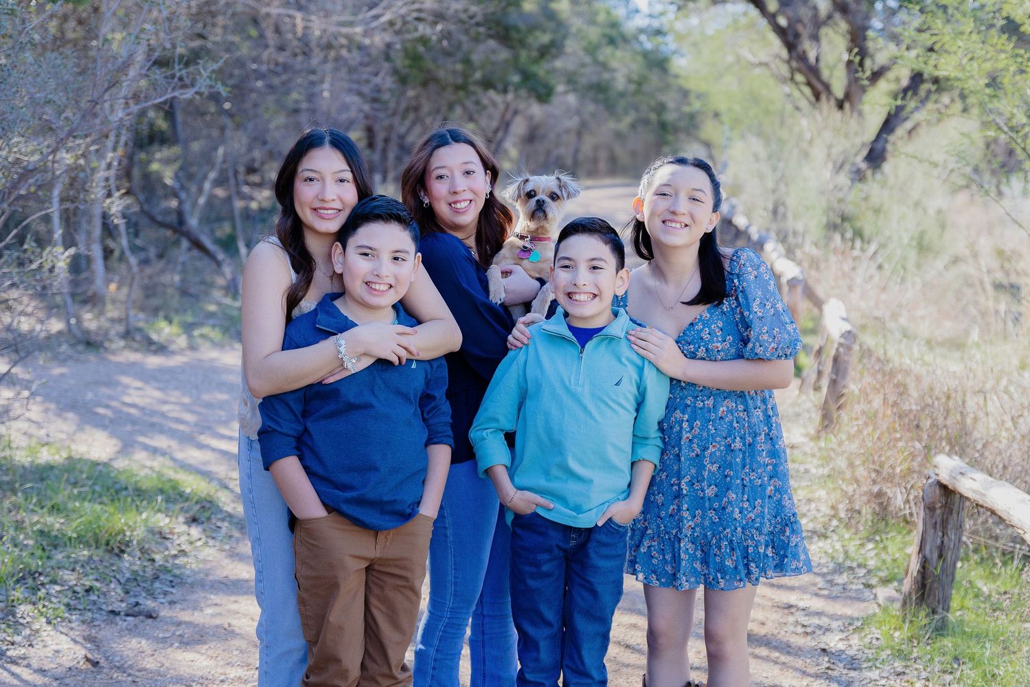 A group poses together smiling on a nature trail with trees and sunlight in the background.