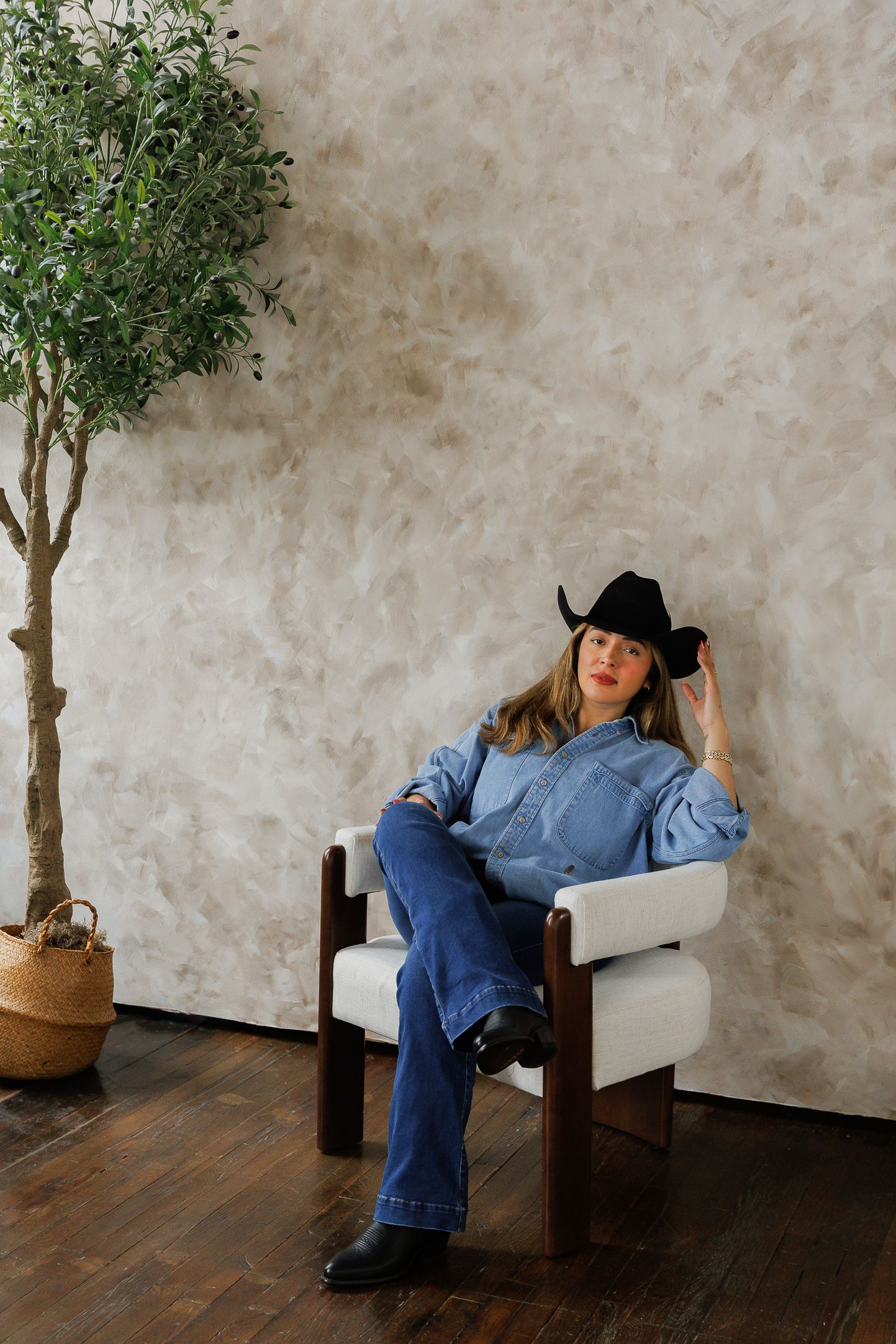 Woman in denim outfit and black cowboy hat sits relaxed in modern white chair beside potted olive tree on hardwood floor.