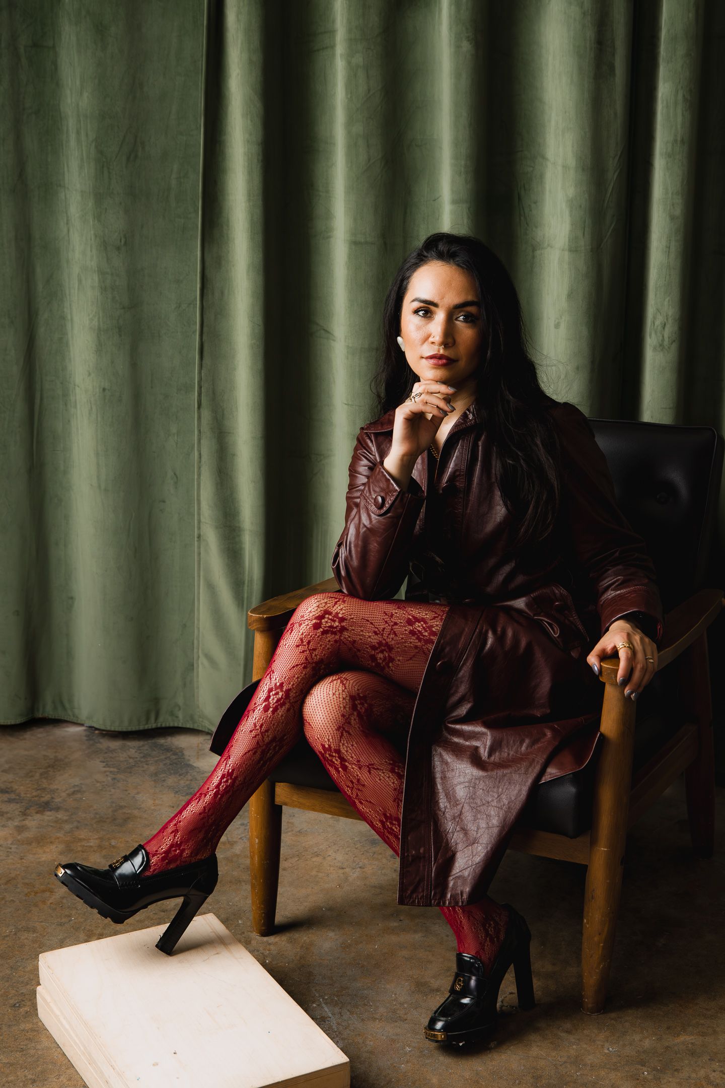 Woman in burgundy leather coat and red lace tights seated in wooden chair against green velvet curtain.