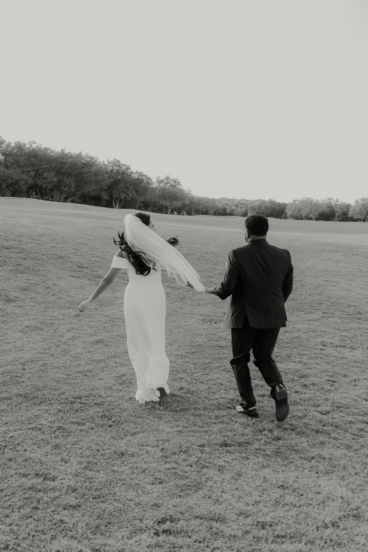 Black and white photo of a couple running playfully across a field after their wedding ceremony.