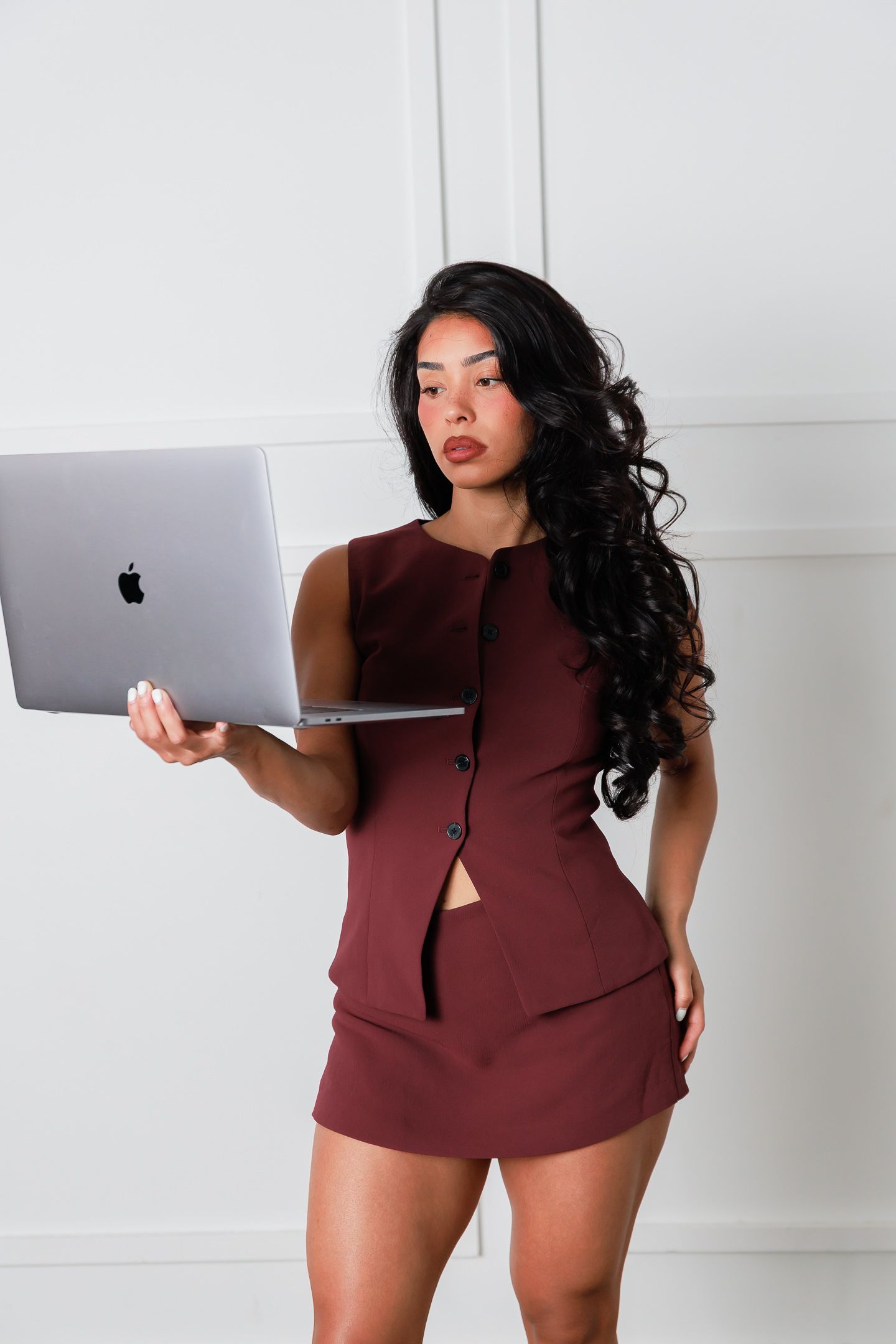 Woman in burgundy skirt suit holding Apple MacBook laptop, posing confidently against white wall background.
