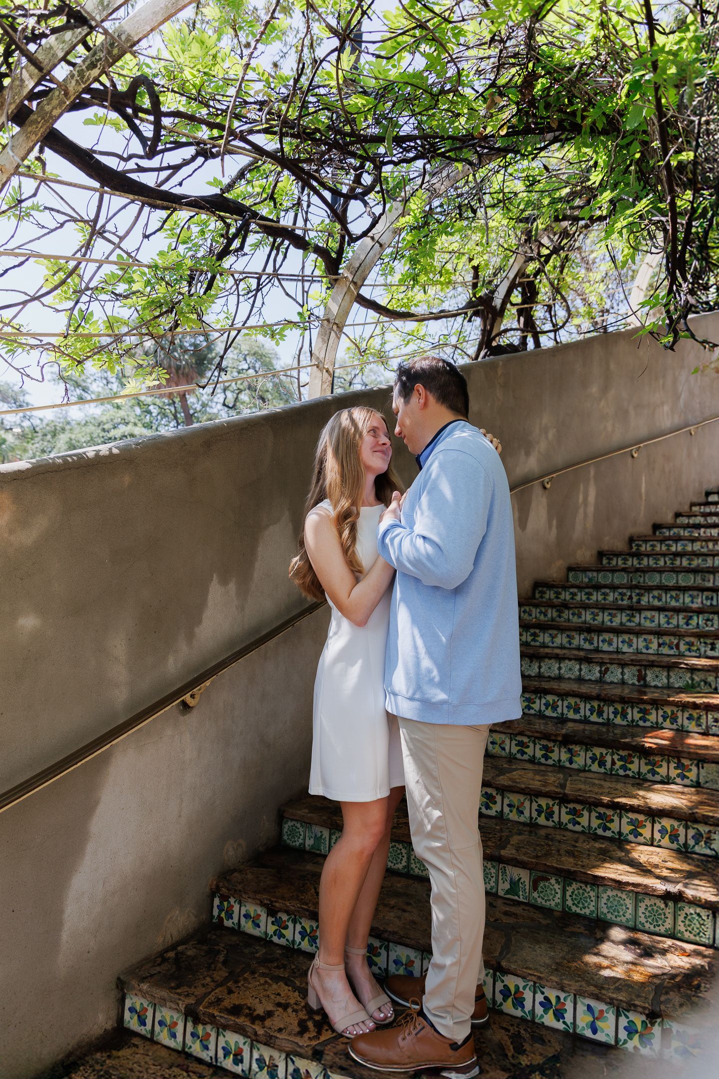 Couple embracing on decorative tiled steps under lush green tree canopy, woman in white dress, man in light blue shirt.