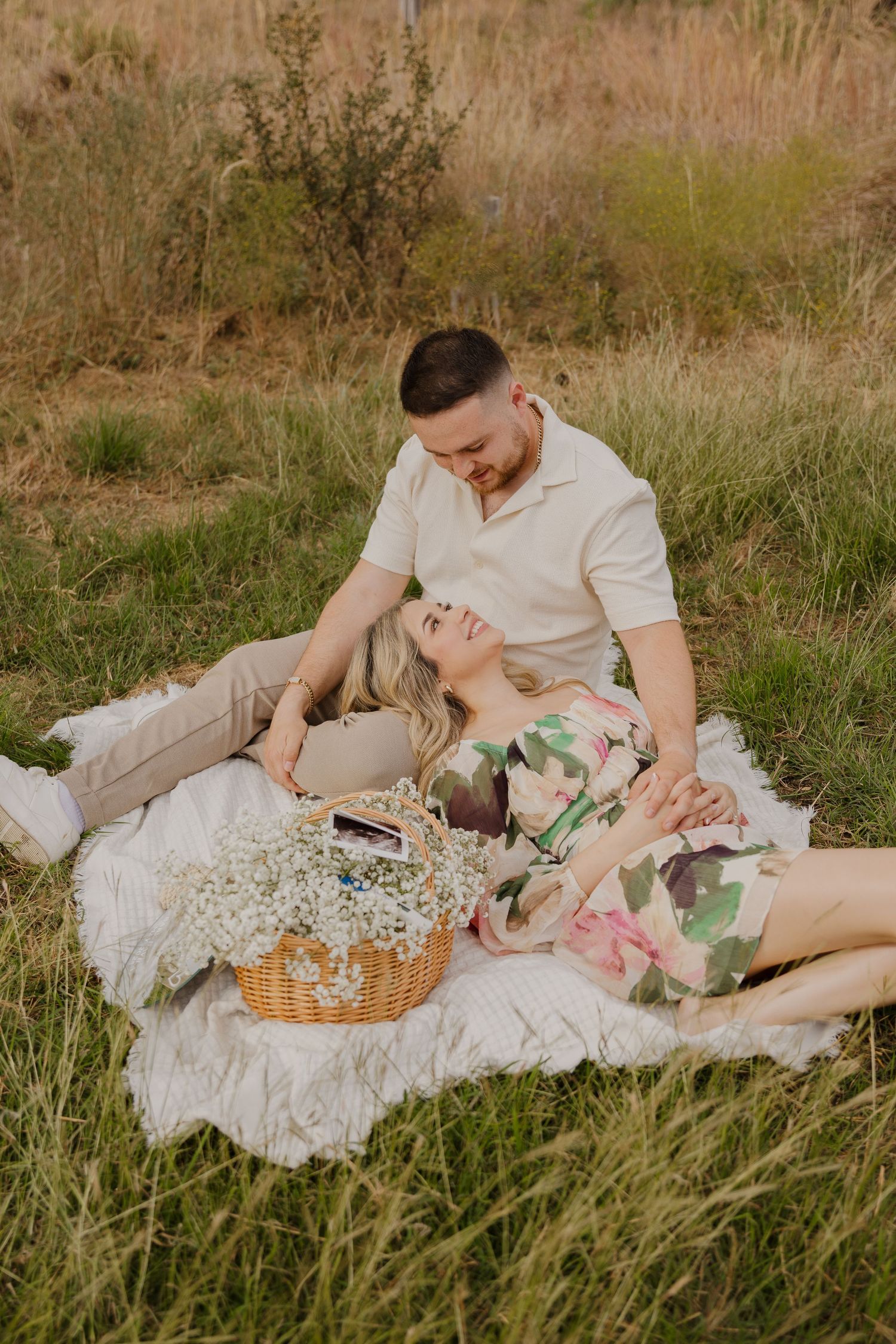 Couple relaxing on picnic blanket with wicker basket in grassy field during photoshoot.