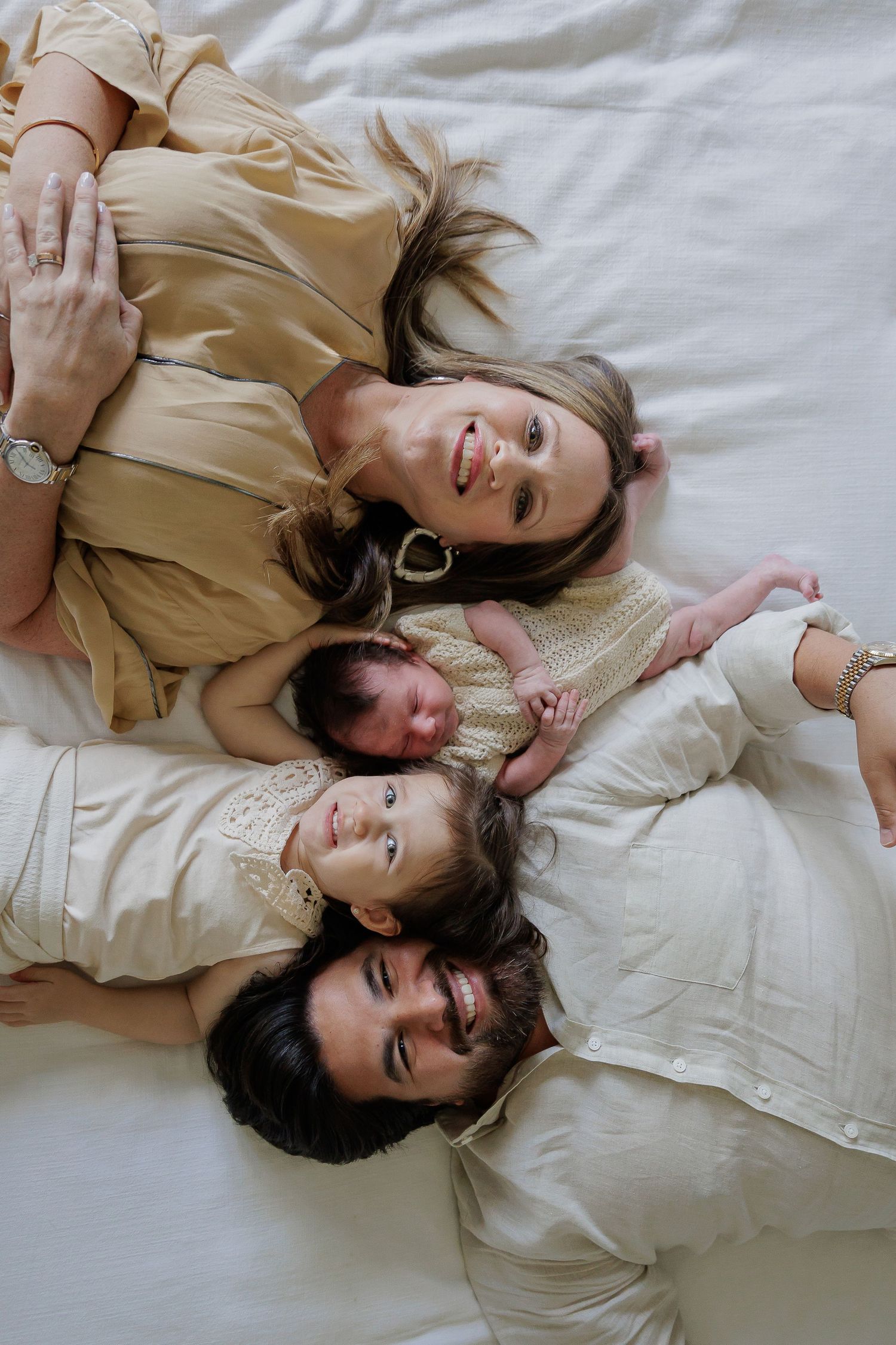 A family in neutral colored clothing lies together on white bedding while smiling at the camera from above.