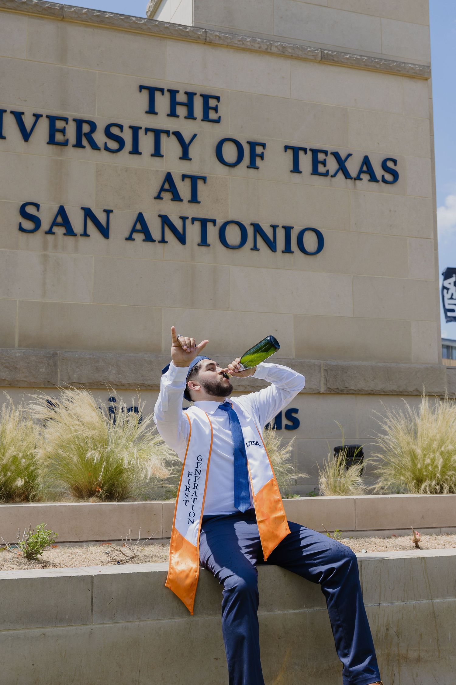 A person in business attire takes a celebratory graduation photo in front of The University of Texas at San Antonio sign.