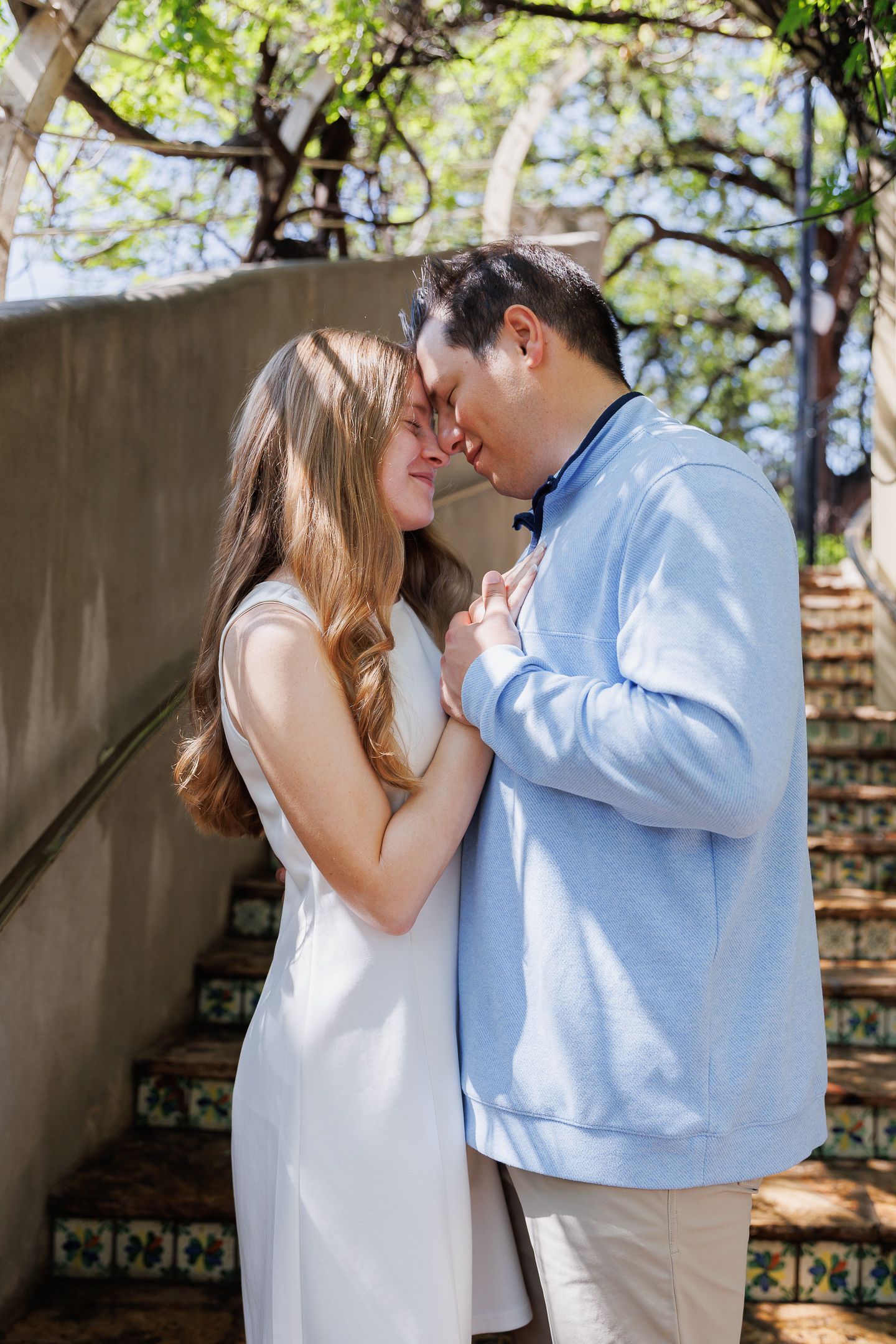 Romantic couple sharing a tender moment outdoors, woman in white dress and man in blue sweater touching foreheads.