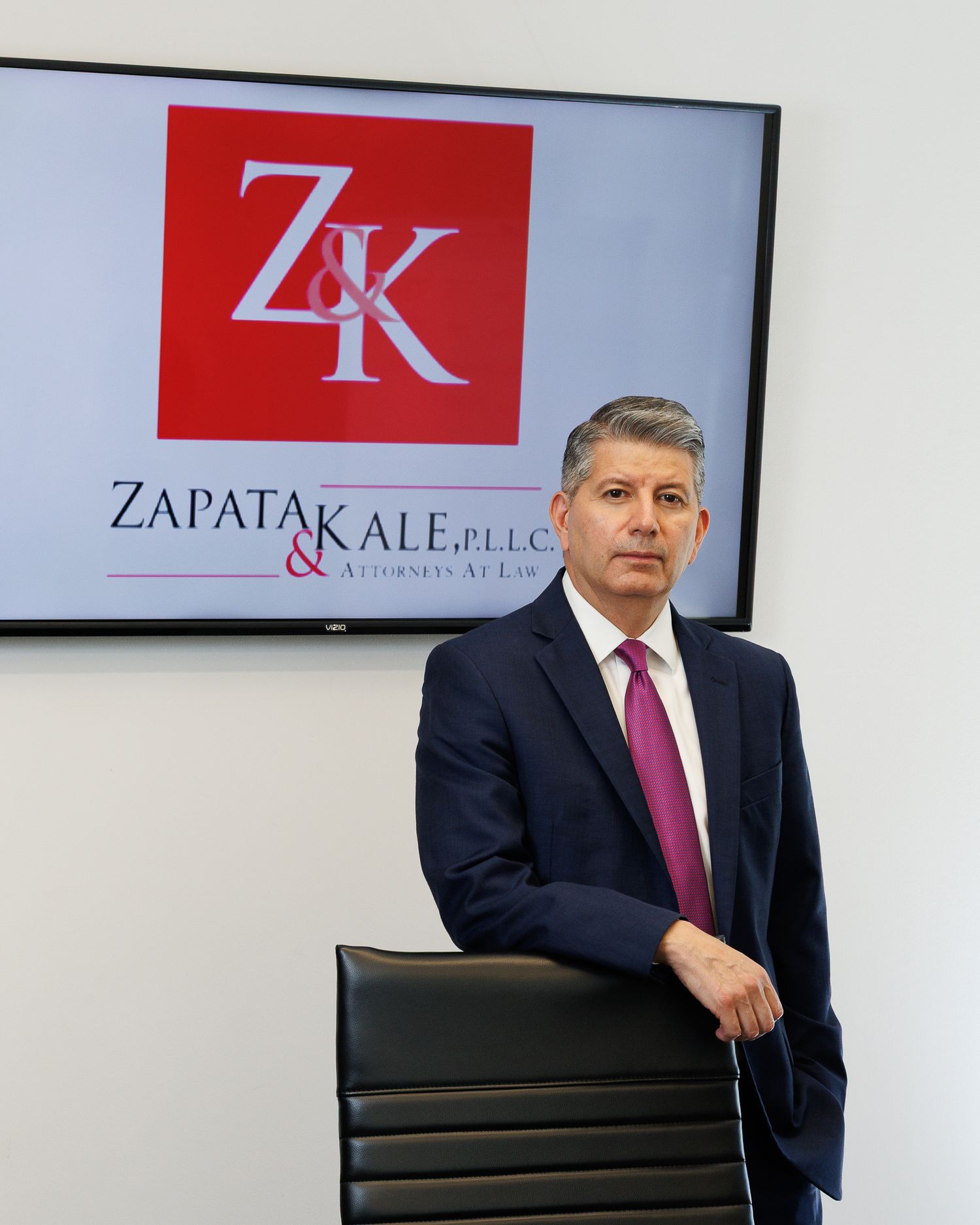 Attorney poses confidently beside Zapata & Kale PLLC law firm sign in professional office setting.