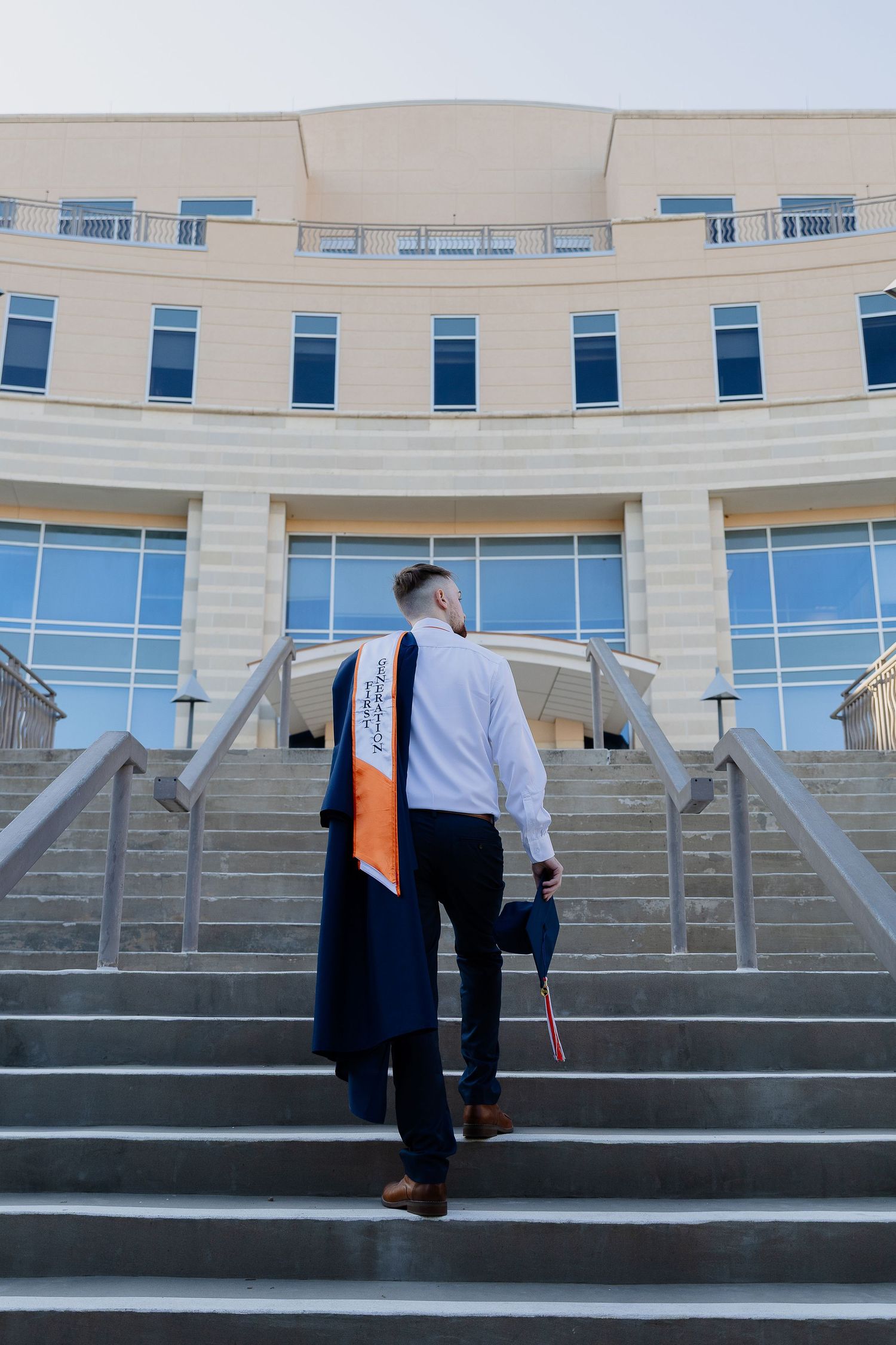 Graduate climbing stairs in front of modern university building wearing graduation stole and holding diploma.