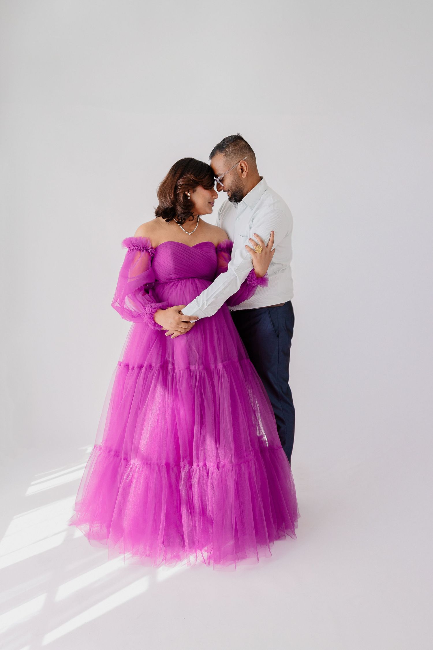 A couple embraces during maternity photos, with the woman wearing a stunning purple tulle gown.