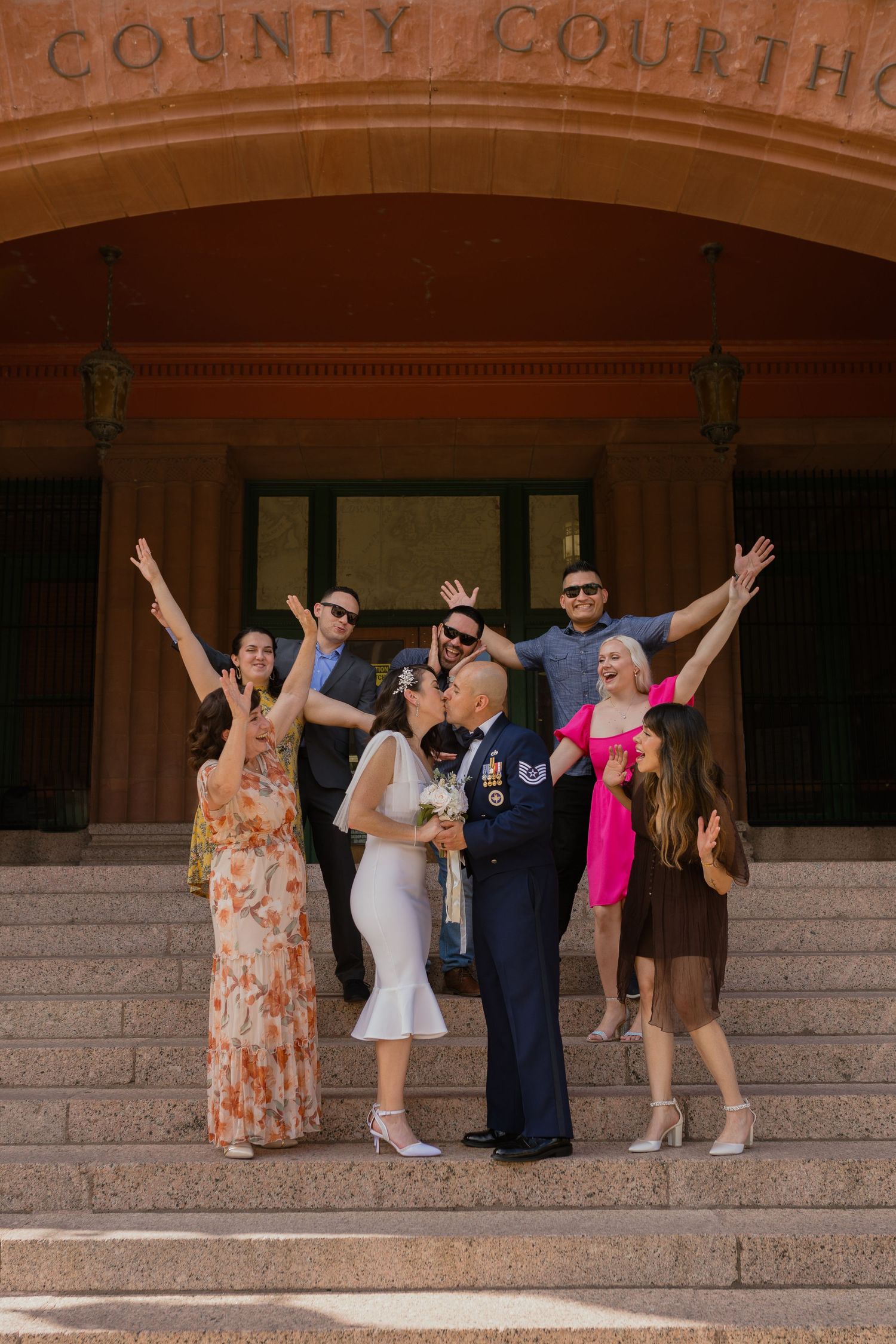 A celebratory group poses joyfully on the steps of a county courthouse after a wedding ceremony.