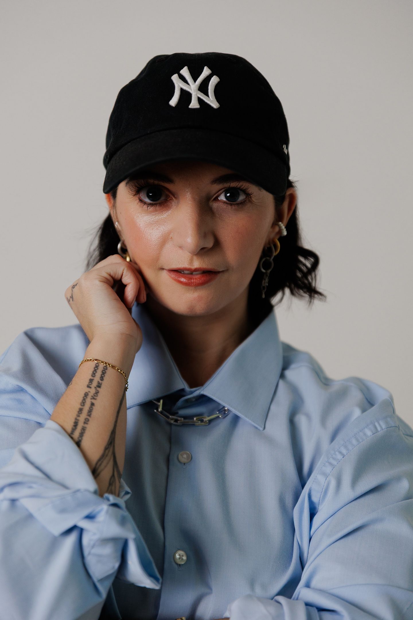 Portrait shot of someone wearing a black NY Yankees baseball cap and light blue button-up shirt with visible arm tattoo.