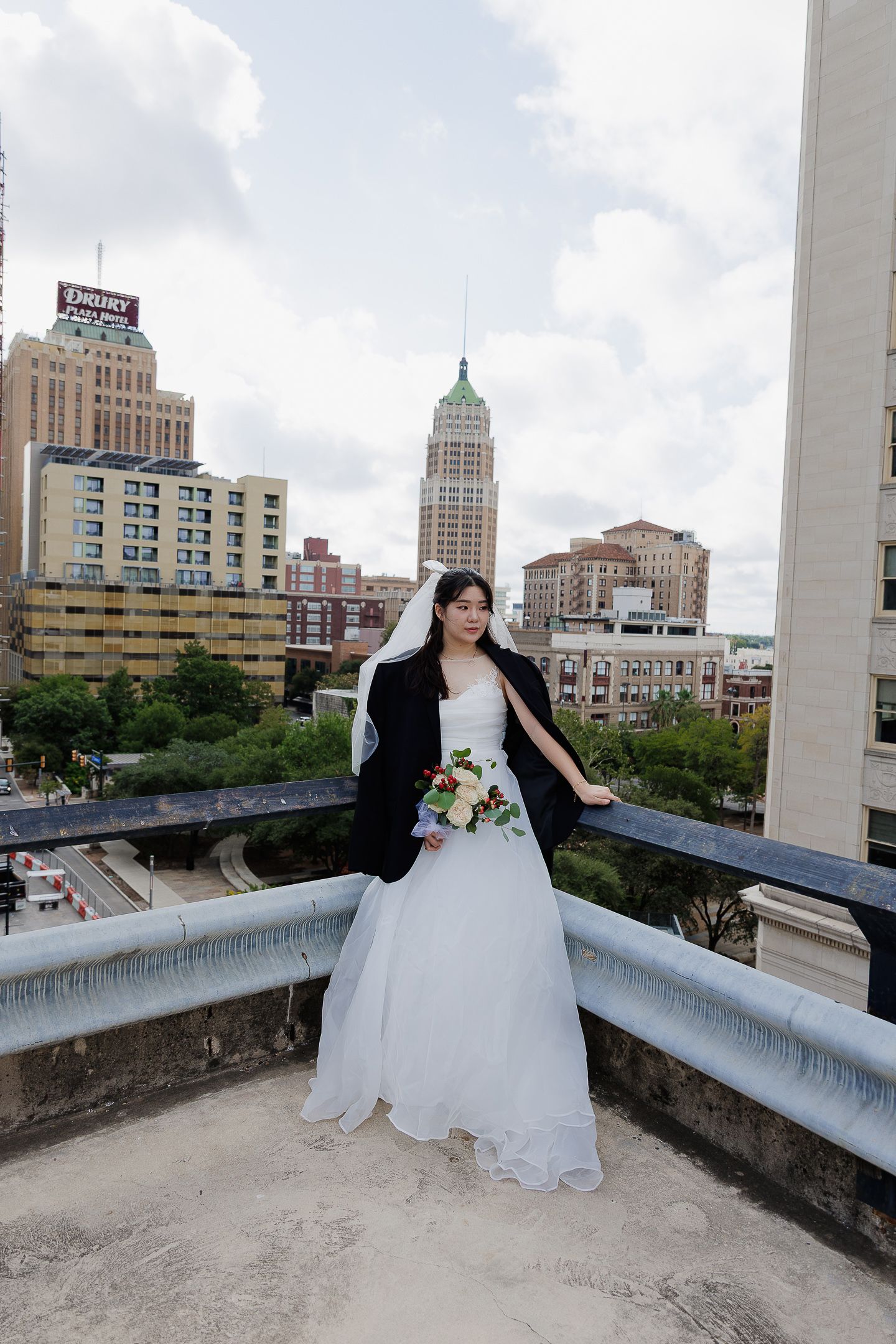A bride in a white ball gown poses on an urban rooftop with city buildings visible in the background.