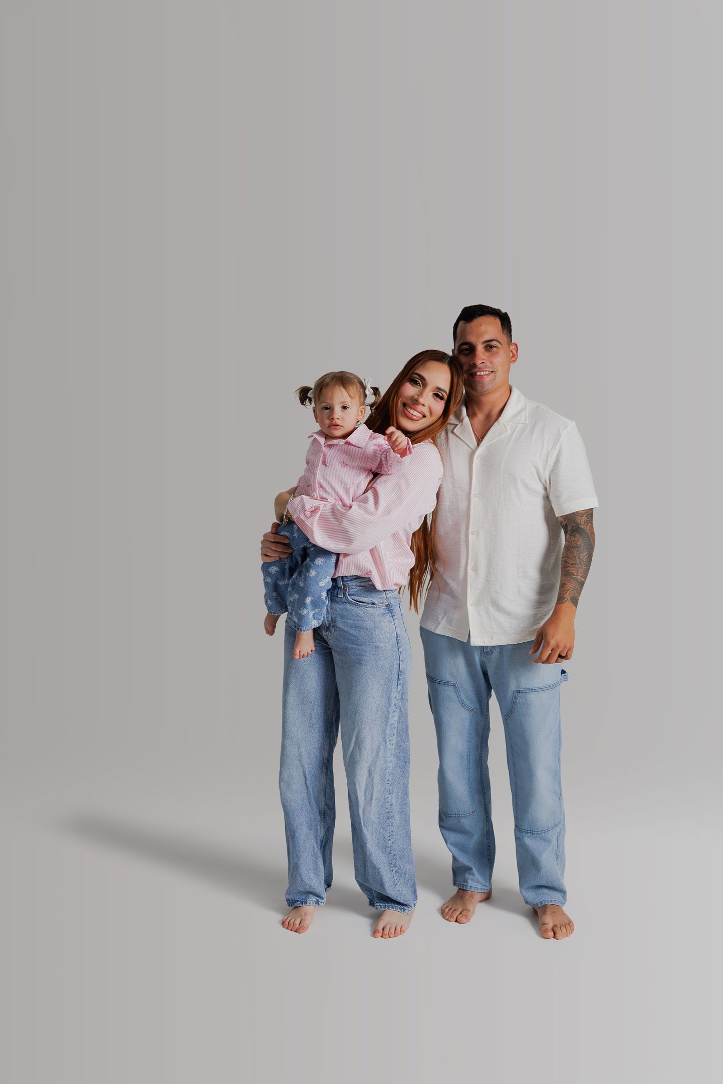 A family in coordinated casual denim outfits poses together barefoot against a light gray studio background.