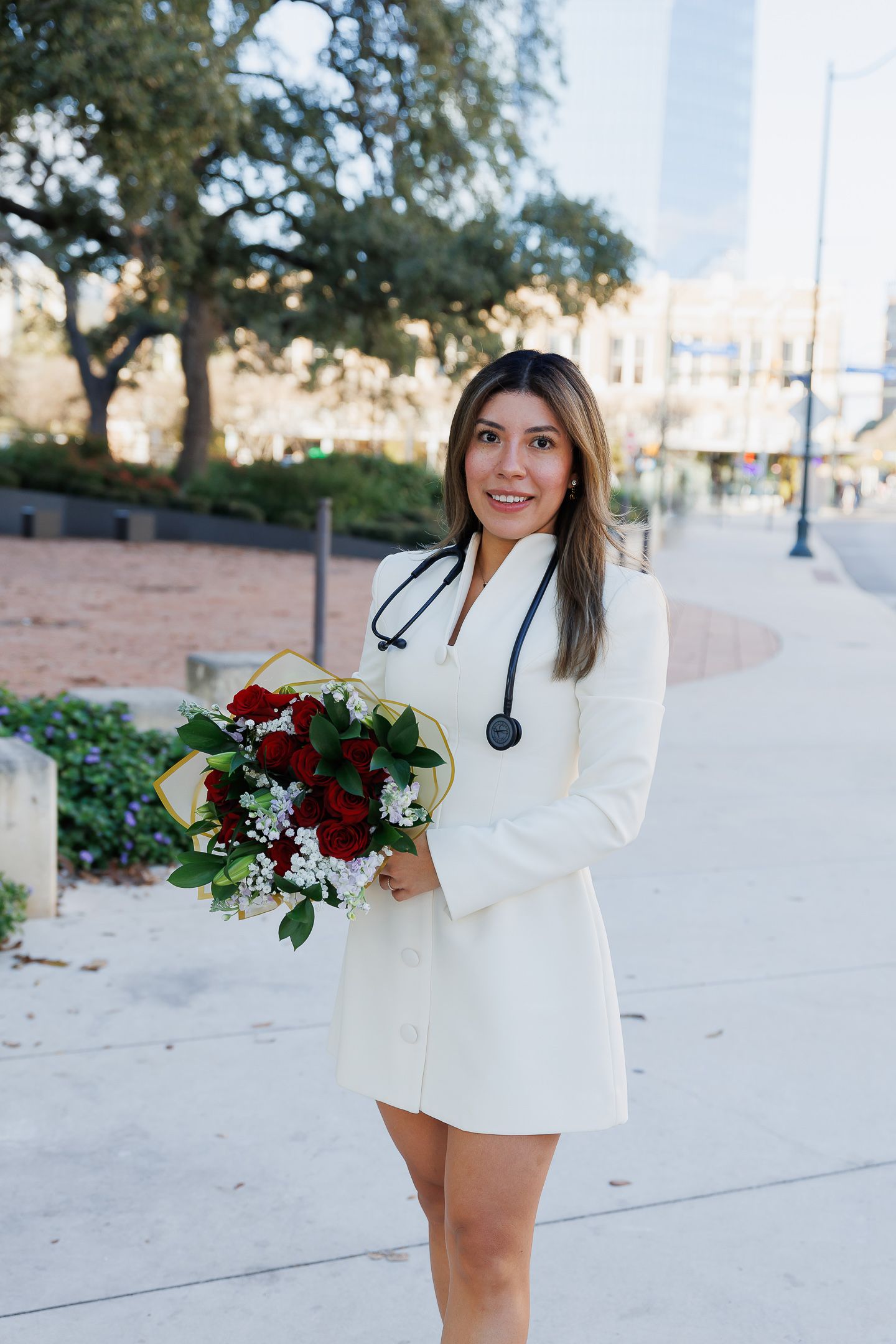 A person in a white blazer holds a bouquet of red and white flowers against a light-colored building exterior.