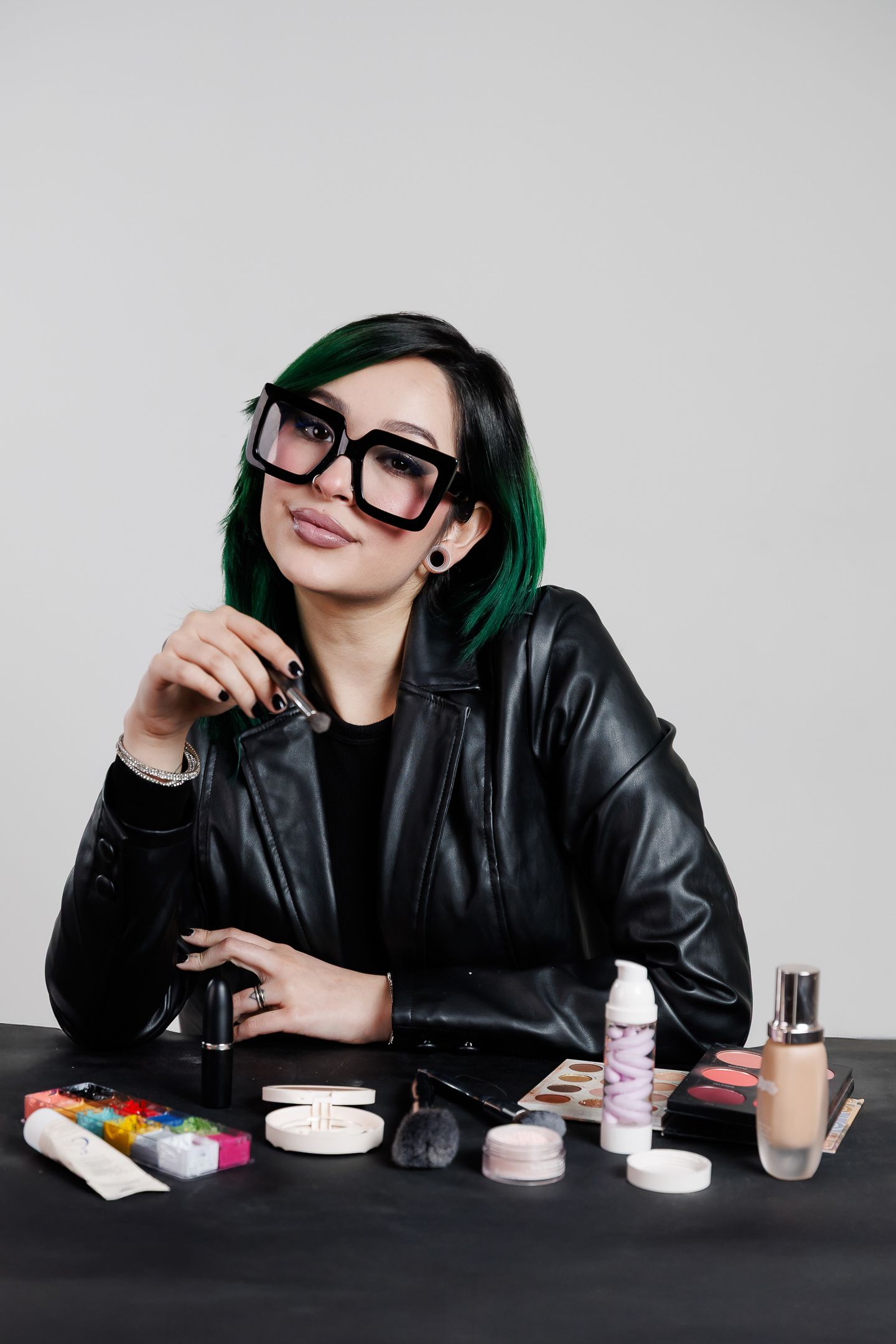 Person with green hair and black leather jacket sits at table with makeup and beauty products arranged in front of them.