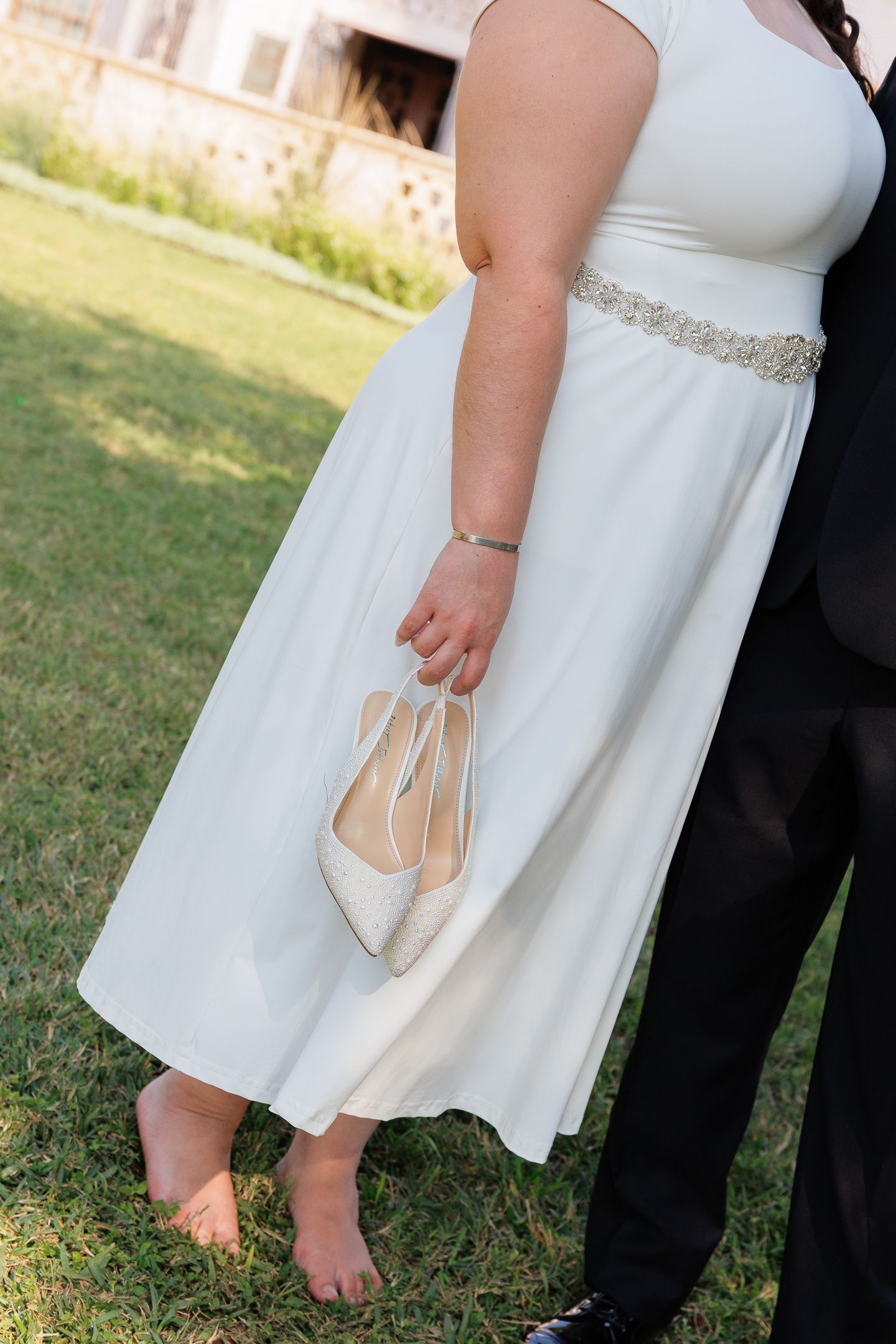A close-up of a bride in a white dress holding ivory wedding shoes while standing barefoot on grass.