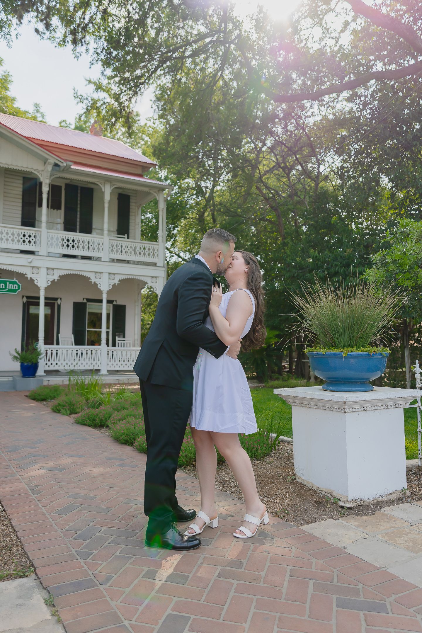 Romantic couple shares intimate moments on a brick pathway outside a charming white colonial-style house.