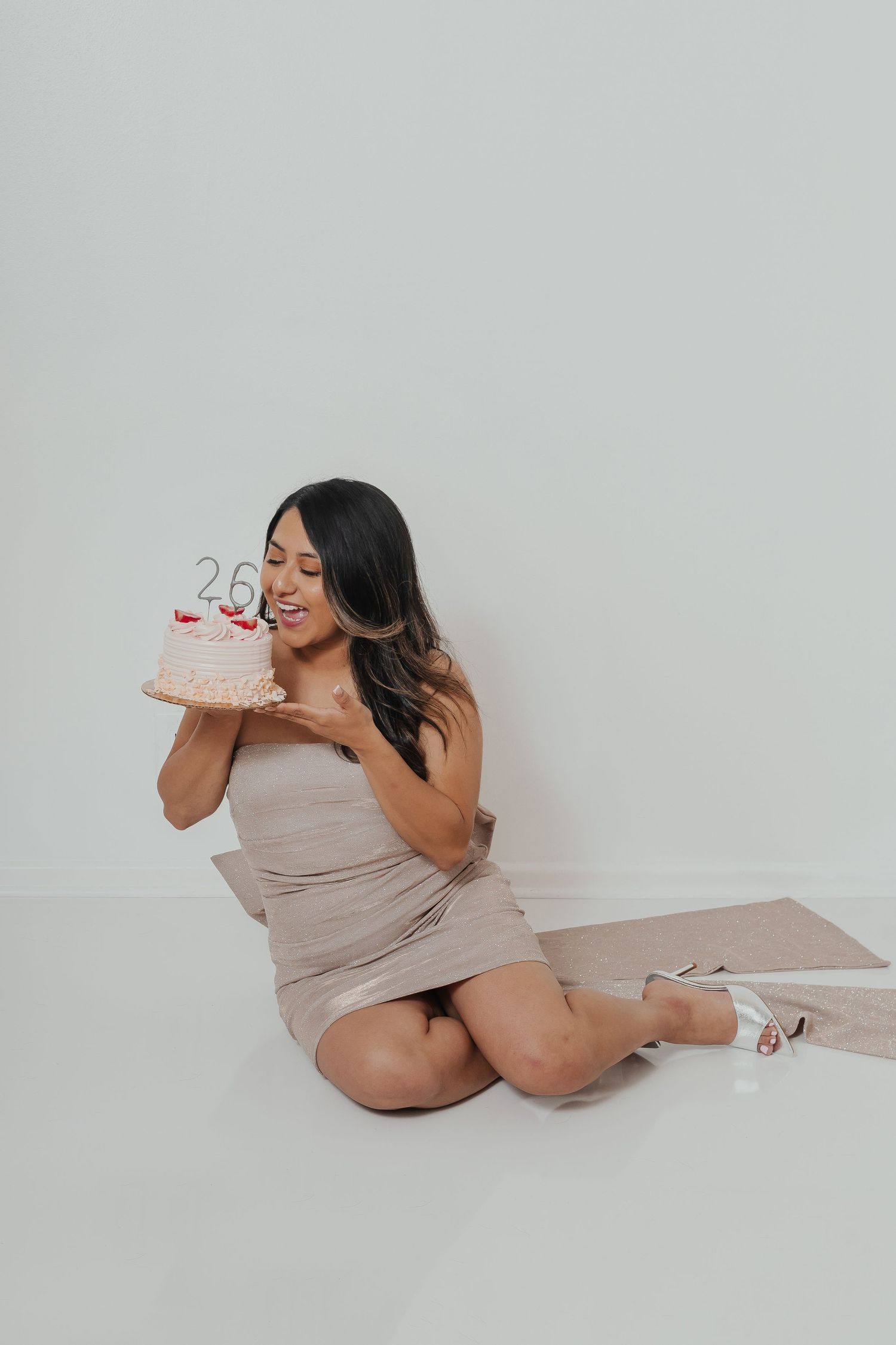 A woman in a beige dress poses candidly with cake against a white backdrop.