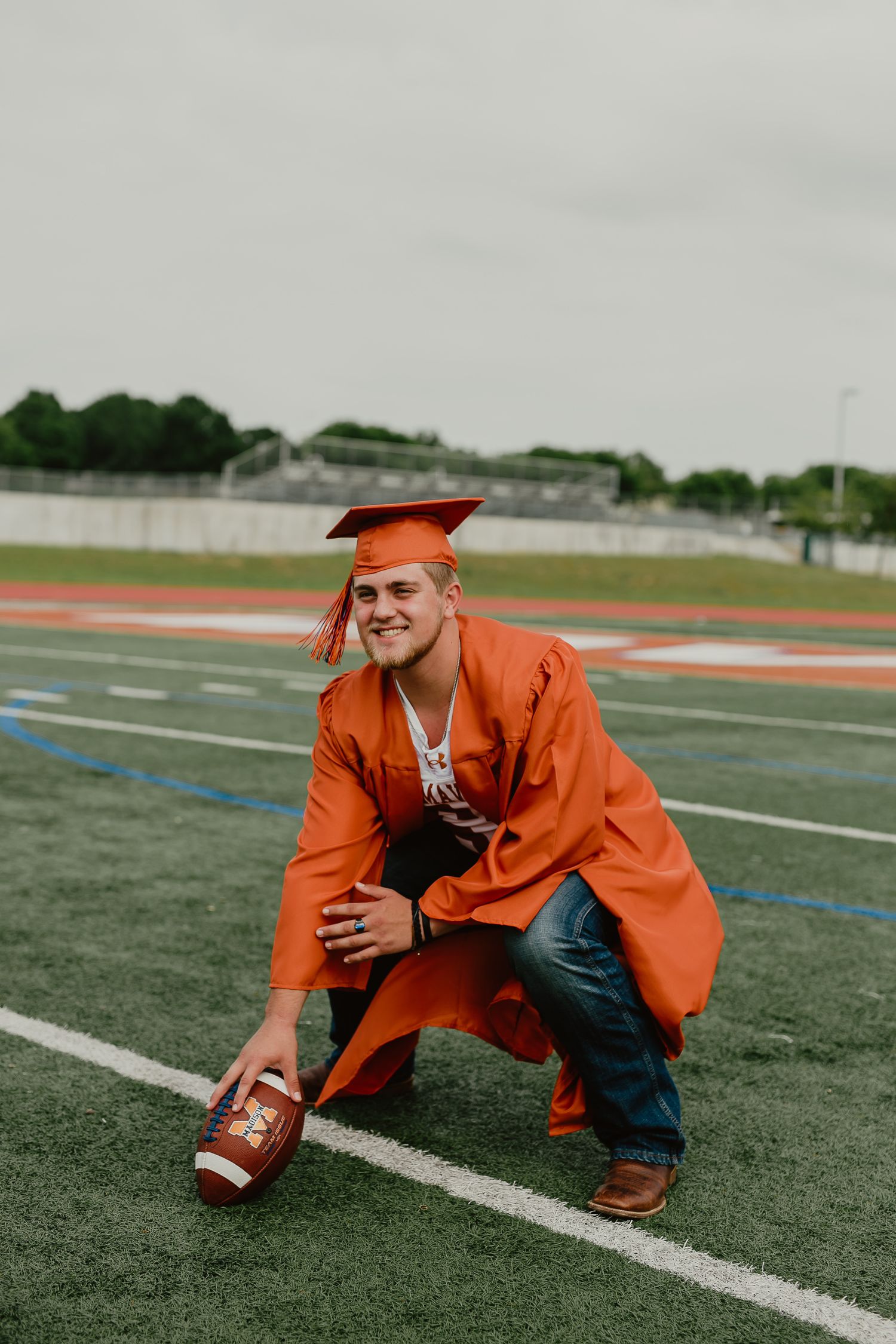 Graduate in orange cap and gown posing with football on sports field.