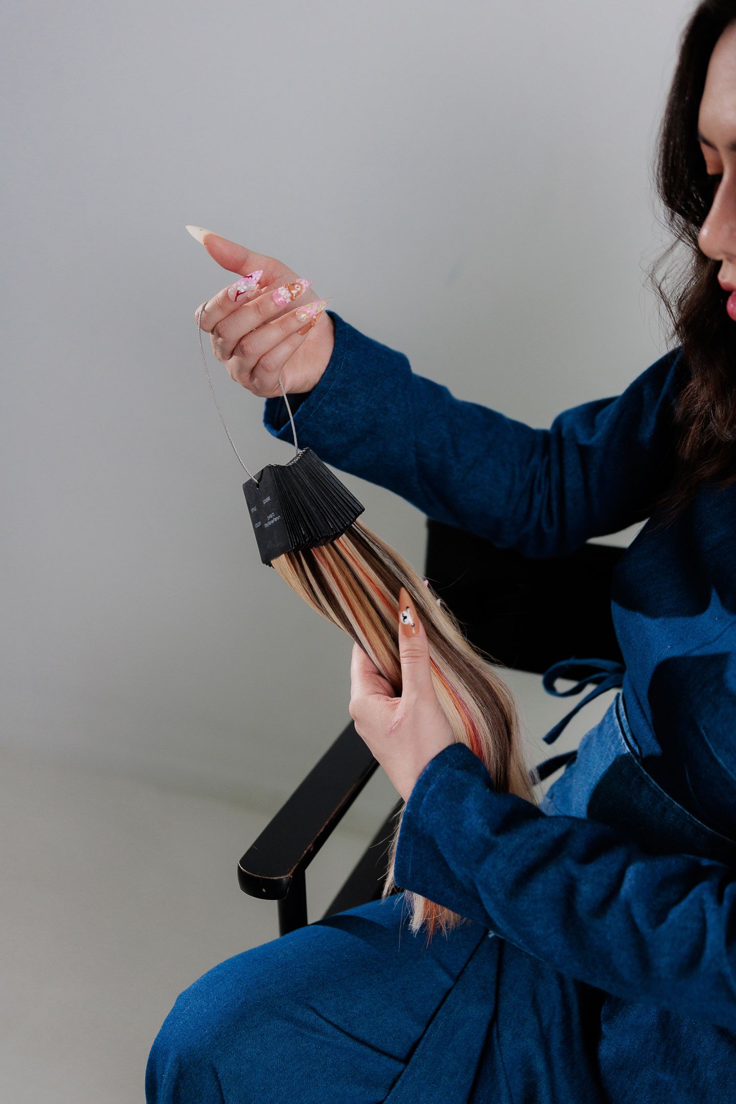Woman in blue denim outfit using a hair straightener on long hair while seated on a black chair.