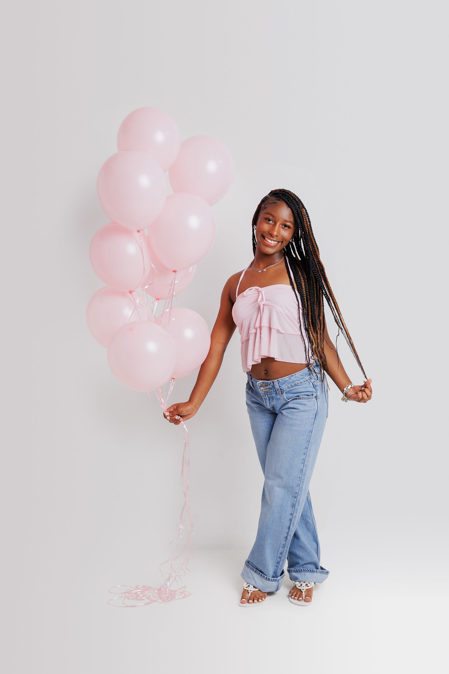 Smiling young woman holding pink balloons, wearing a pink crop top and blue jeans, posing on a white background.