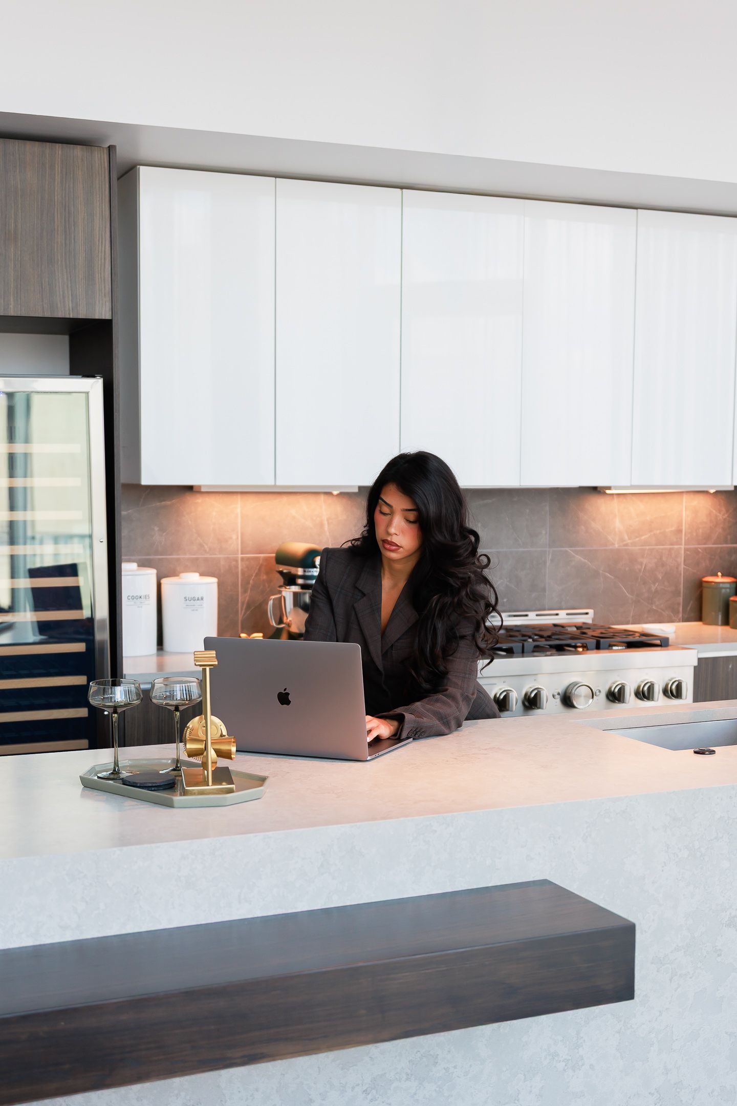 Woman in blazer working on MacBook laptop at modern kitchen counter with white cabinets.