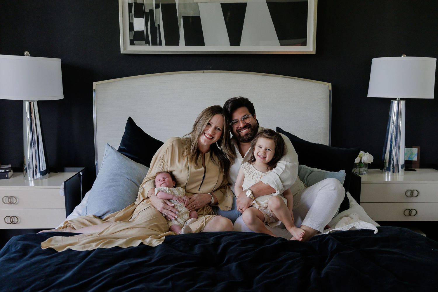 Family snuggling together on white bedding in modern bedroom with dark walls and framed artwork.