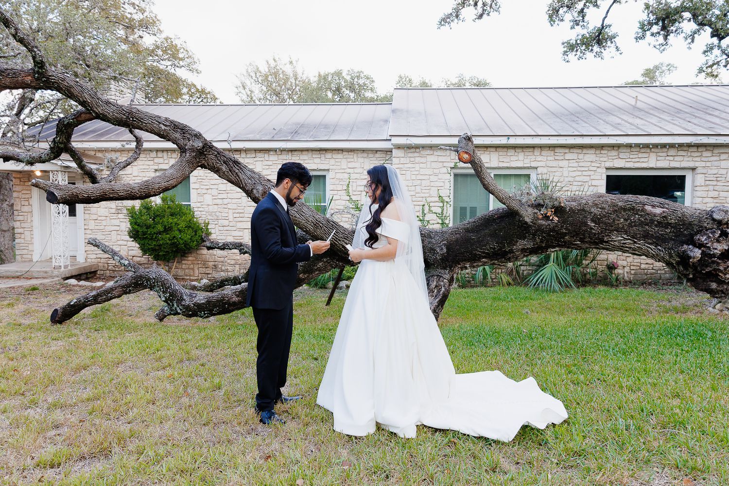 A couple in wedding attire stands together under a large twisted tree branch on a lawn in front of a stone building.