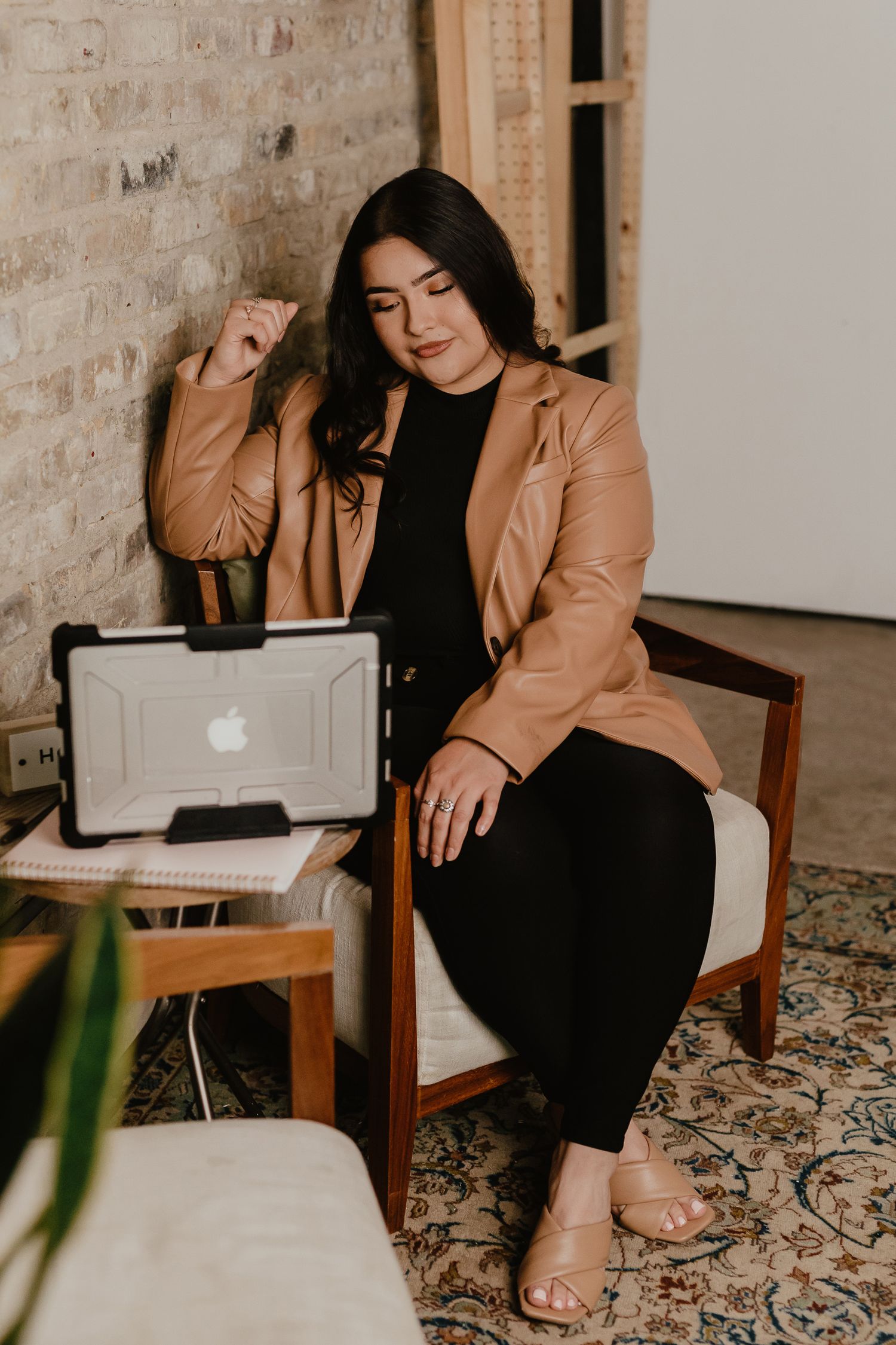Professional in tan blazer works on laptop in modern brick-walled office space.