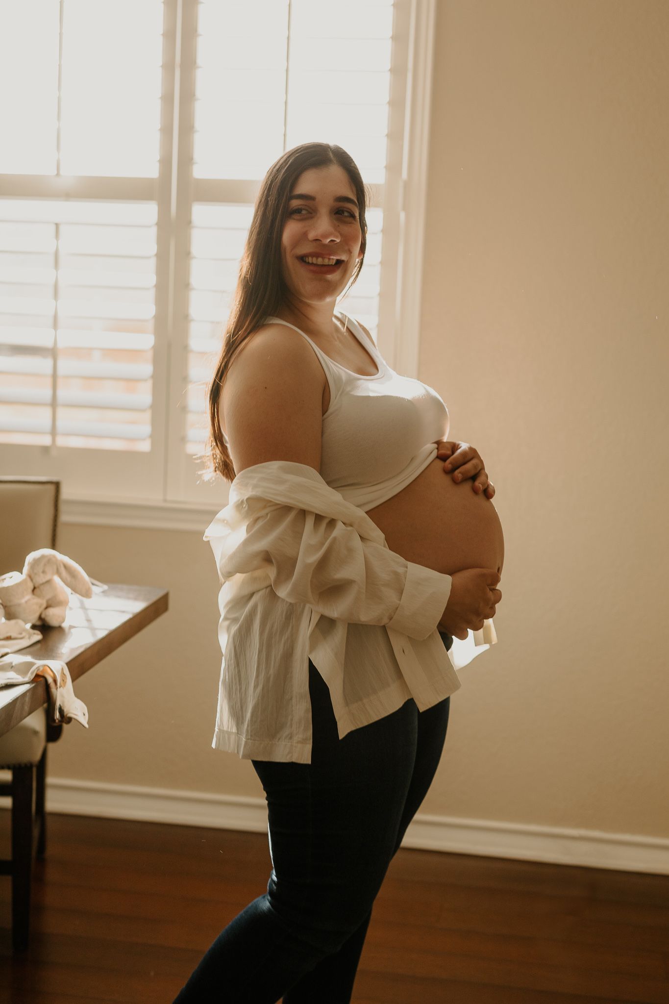 An expecting mother in white top poses by window during maternity photoshoot.
