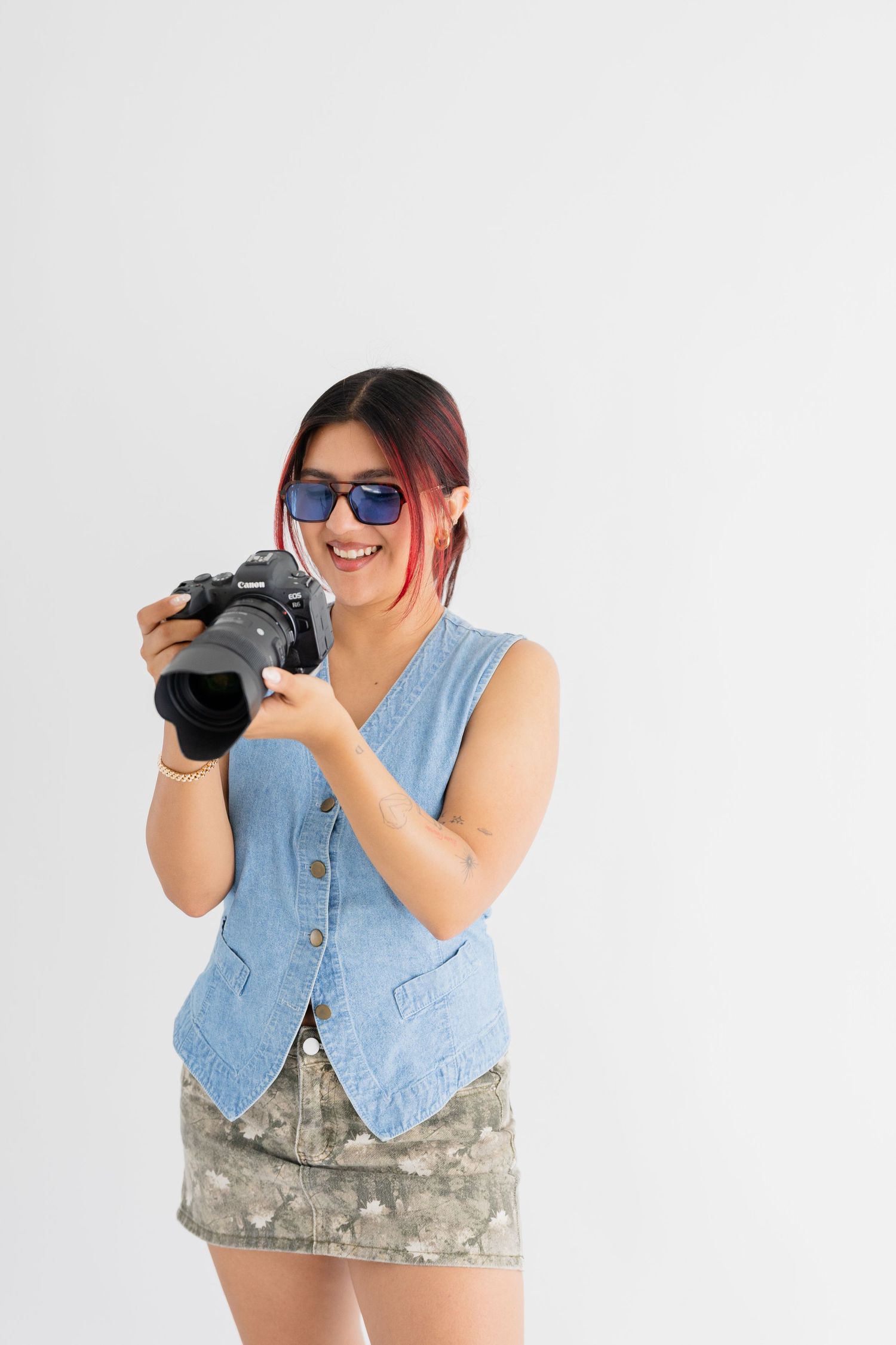 Photographer wearing denim vest and sunglasses holds DSLR camera while smiling against white background.