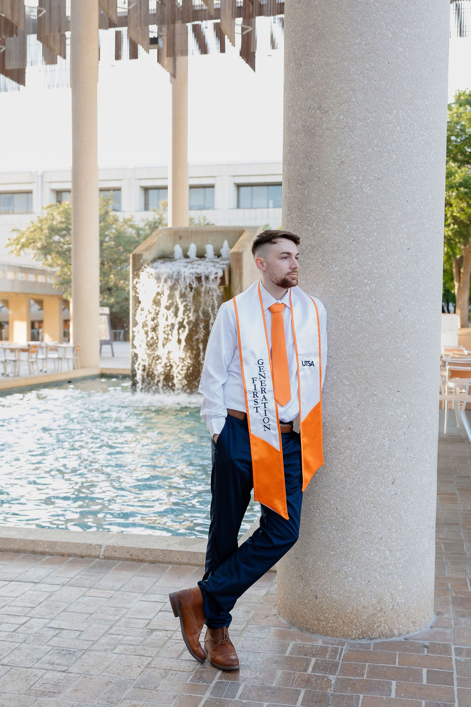 Graduate in navy suit and orange stole stands by fountain column in graduation attire.