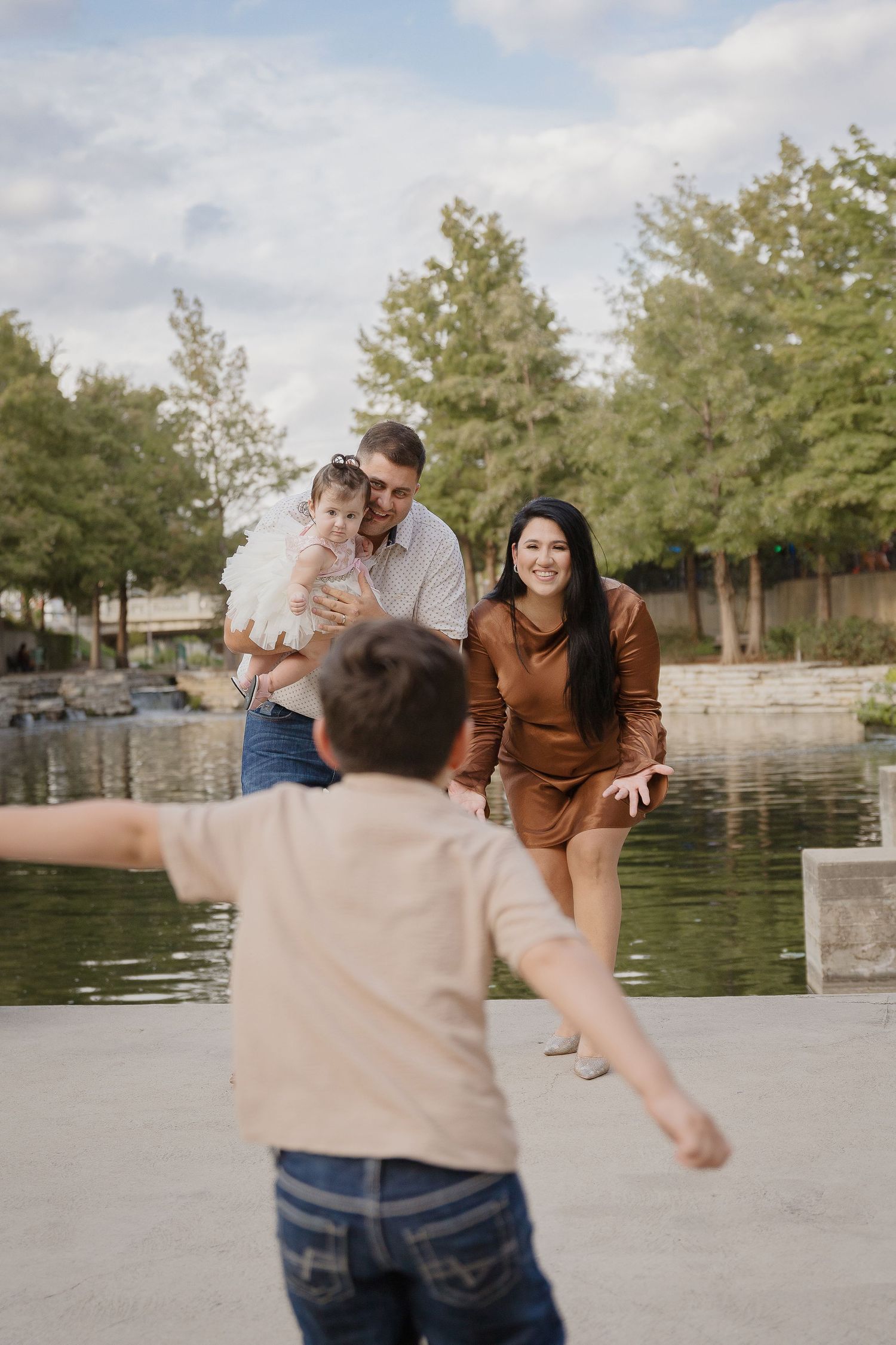 A heartwarming family moment captured by a pond as a young child runs towards loved ones standing near the water's edge.