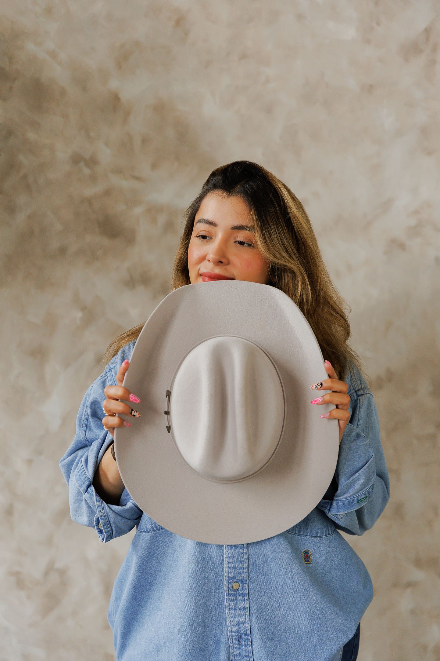 Woman in denim shirt holding a beige cowboy hat in front of her face, smiling, with long hair.