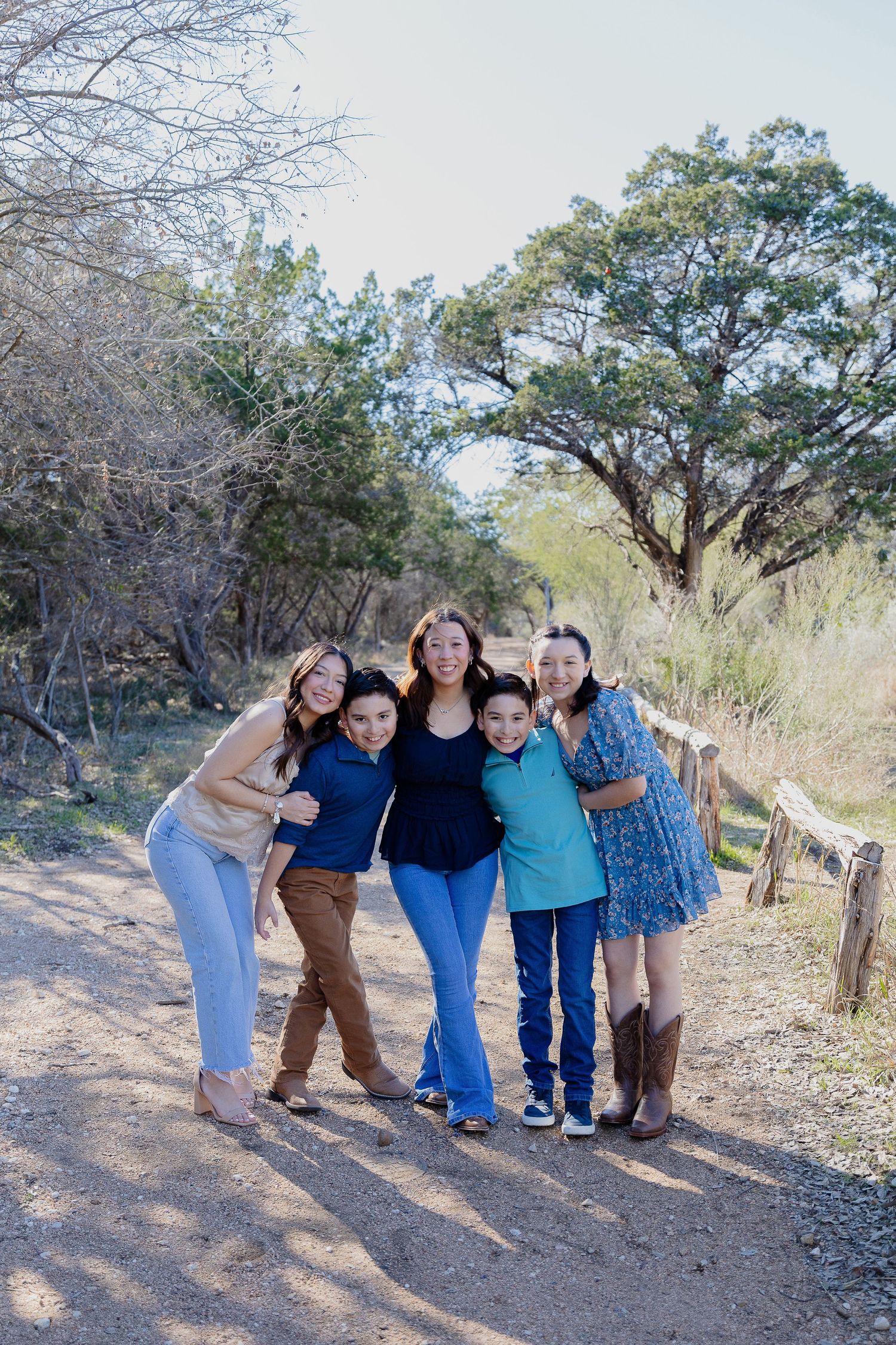 A group poses together smiling on a scenic dirt trail surrounded by trees and natural brush on a sunny day.