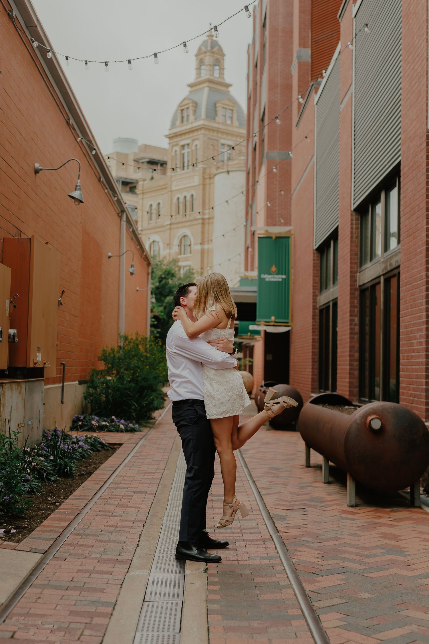 A romantic couple embraces in an urban alley with string lights and a historic domed building visible behind them.