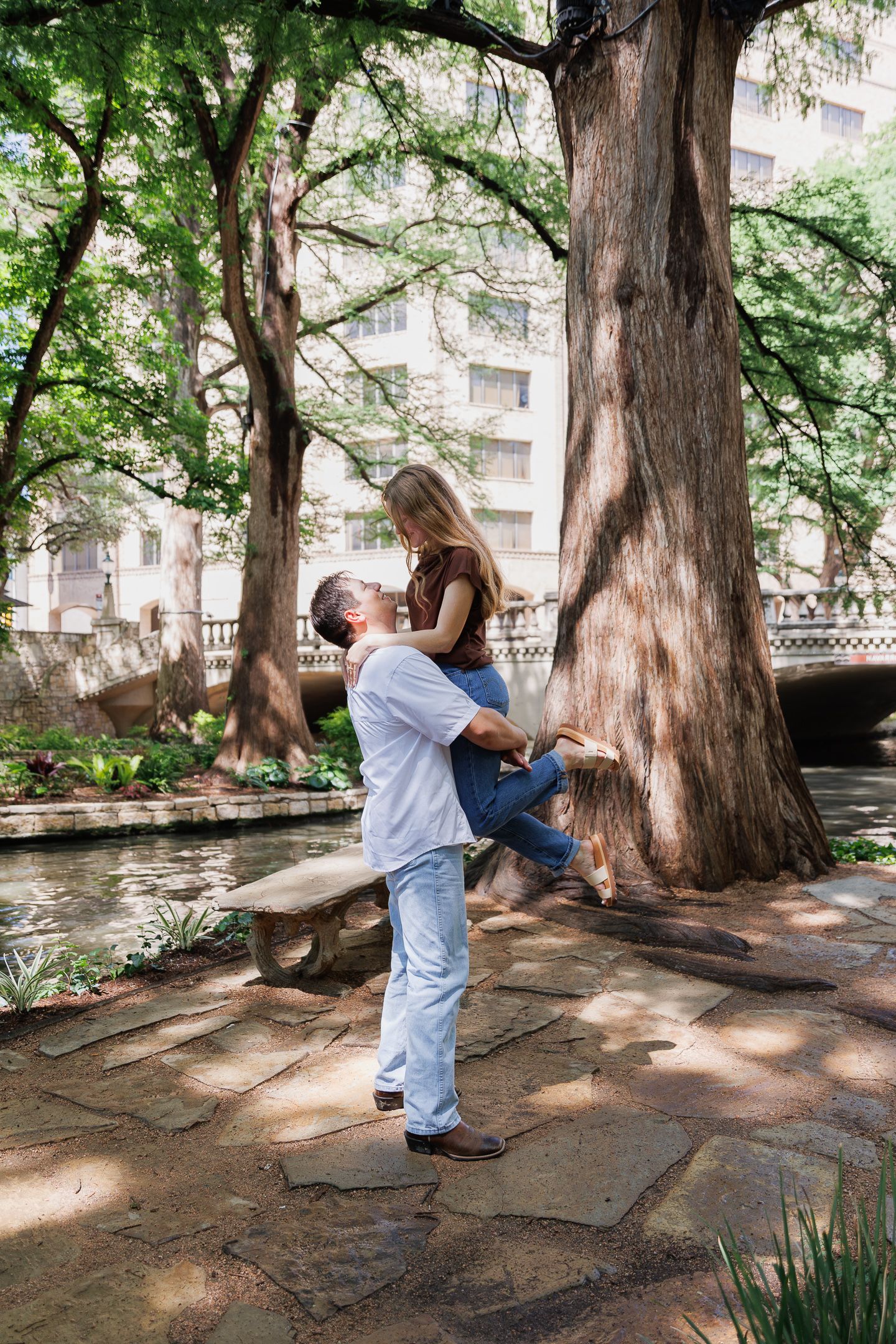 Man lifting woman in his arms outdoors near a scenic river walk surrounded by large trees.