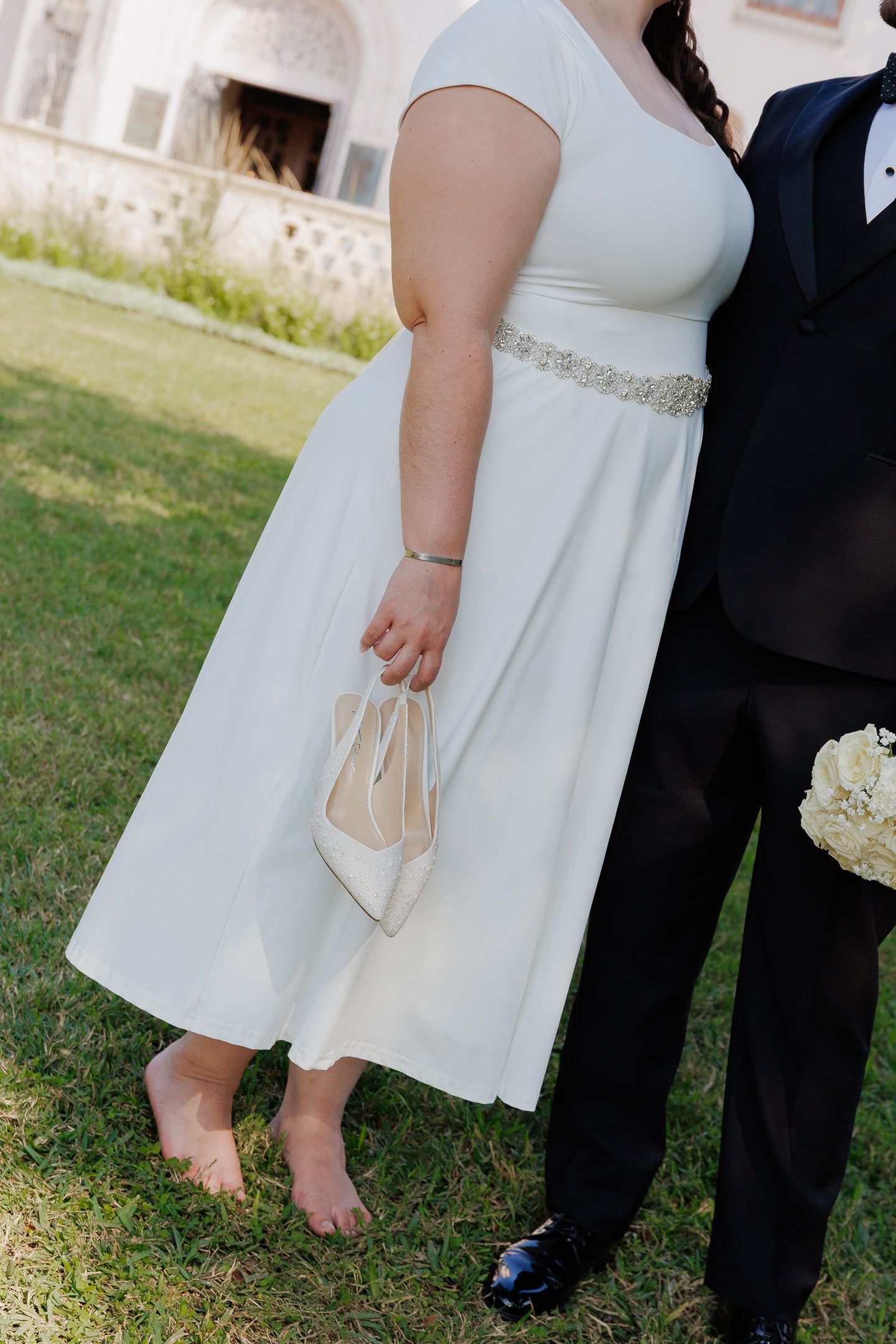 A person in a white dress holds elegant cream-colored heels while standing barefoot on grass next to formal attire.