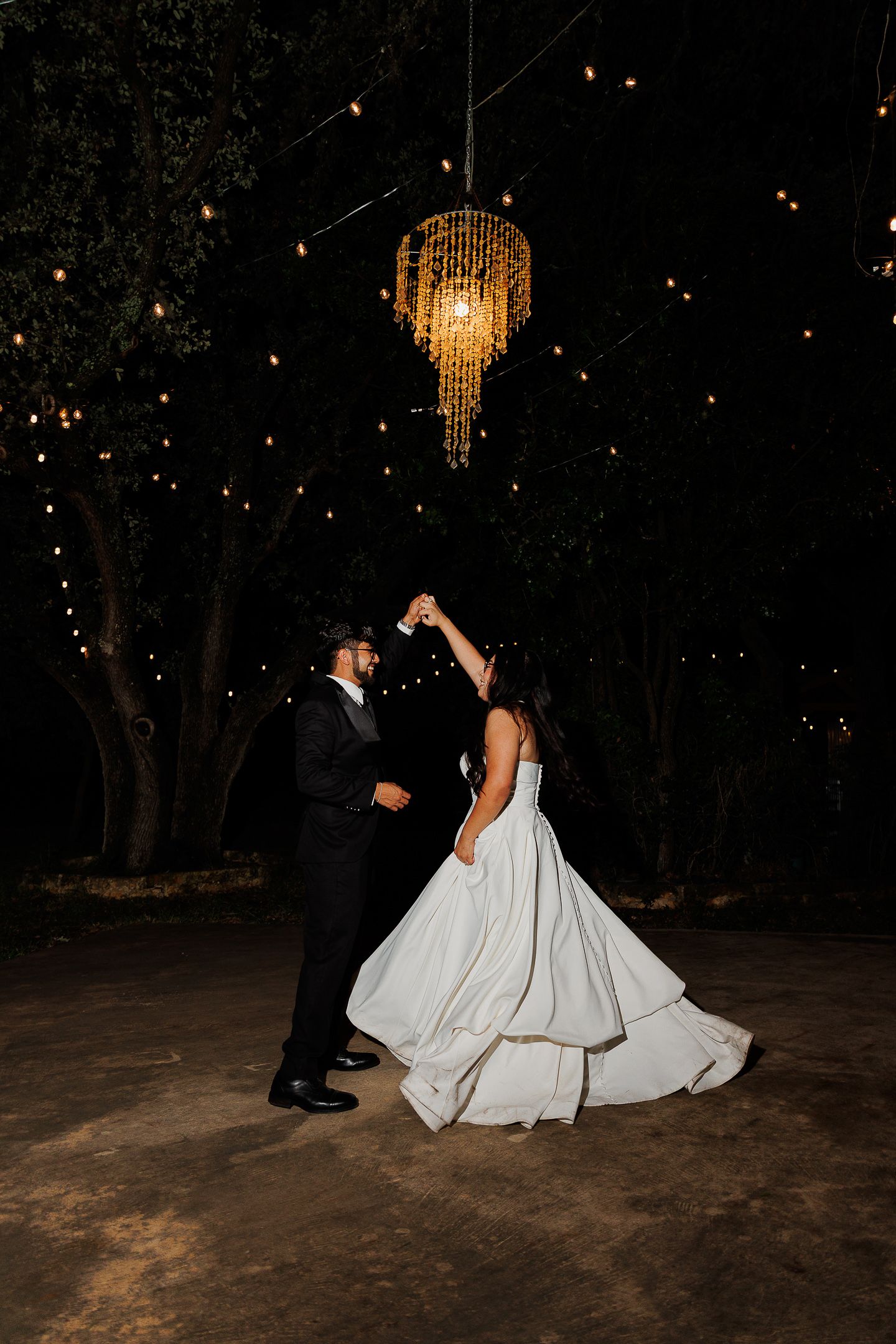 A couple in formal wedding attire dances together under twinkling lights and a crystal chandelier at an outdoor evening reception.