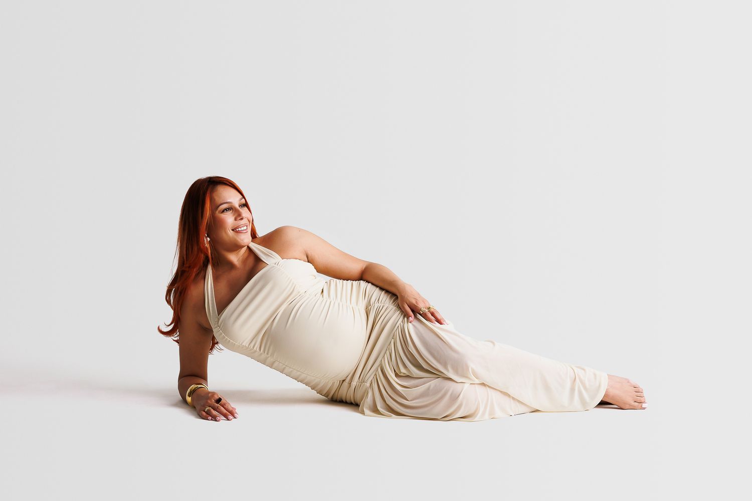A pregnant woman in a cream-colored dress reclines on a white floor during a maternity photoshoot.