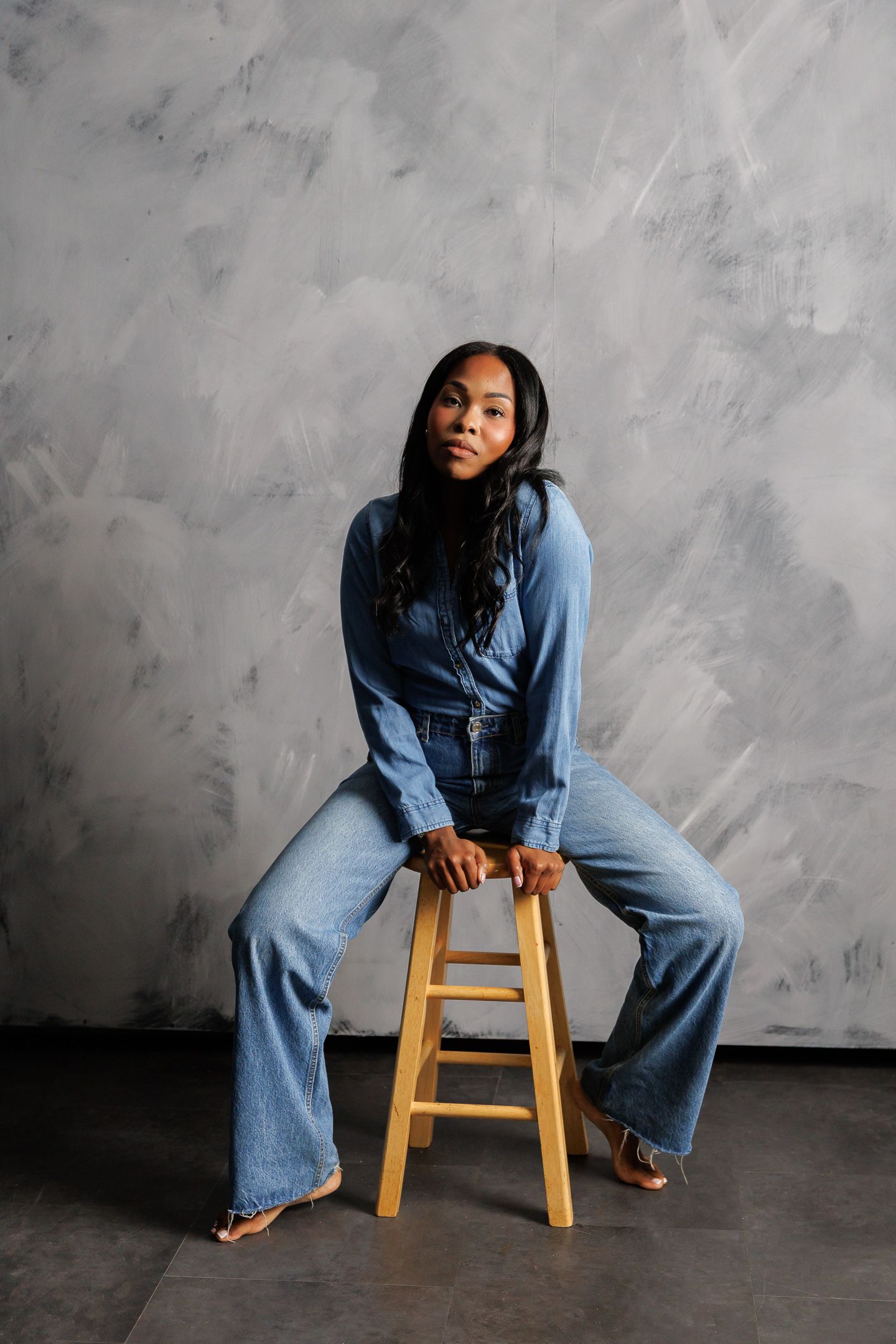 A Black woman in a denim jumpsuit and wide-leg jeans sits on a wooden stool against a gray textured wall.