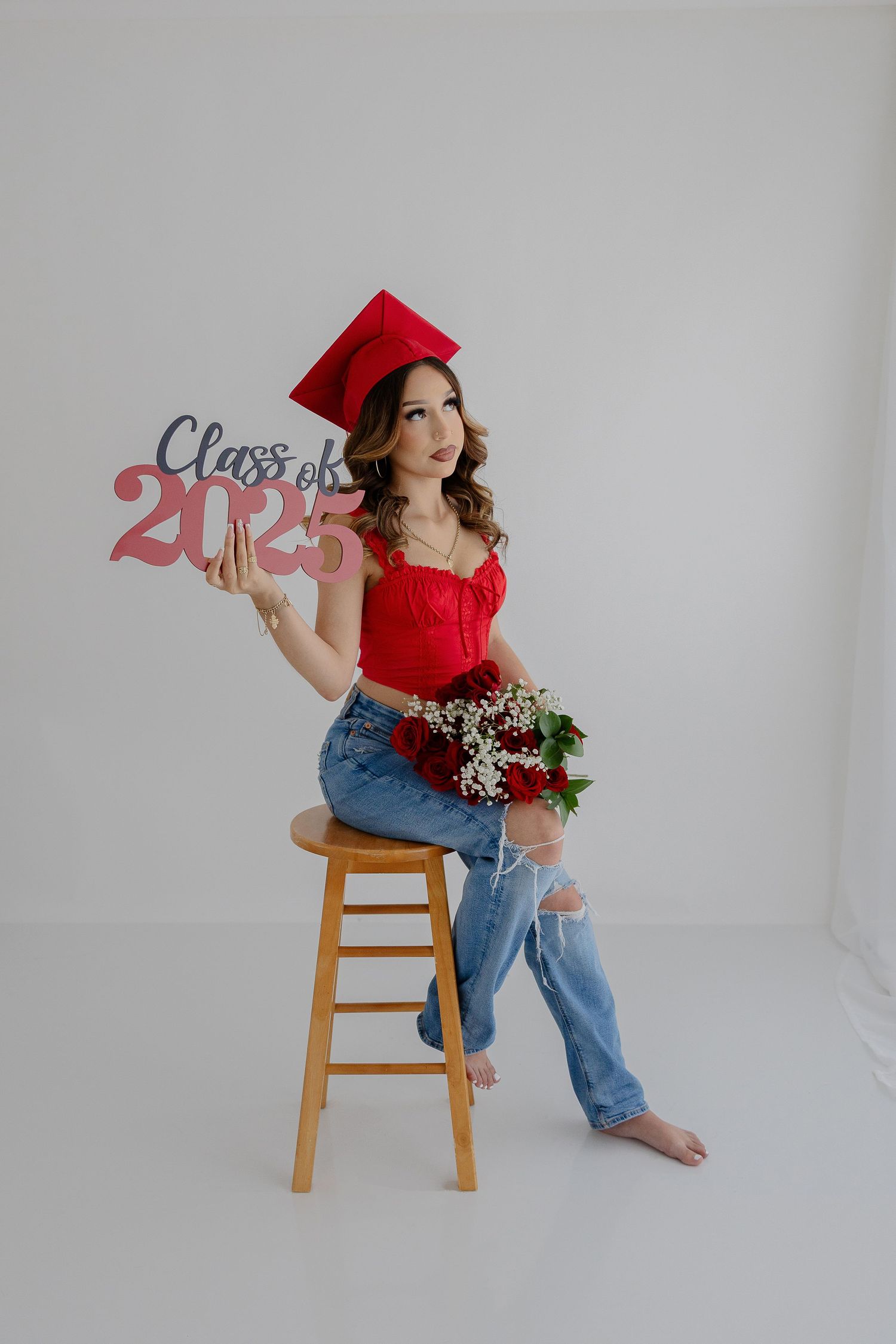 Graduate poses on stool with Class of 2025 sign in red top and distressed jeans.