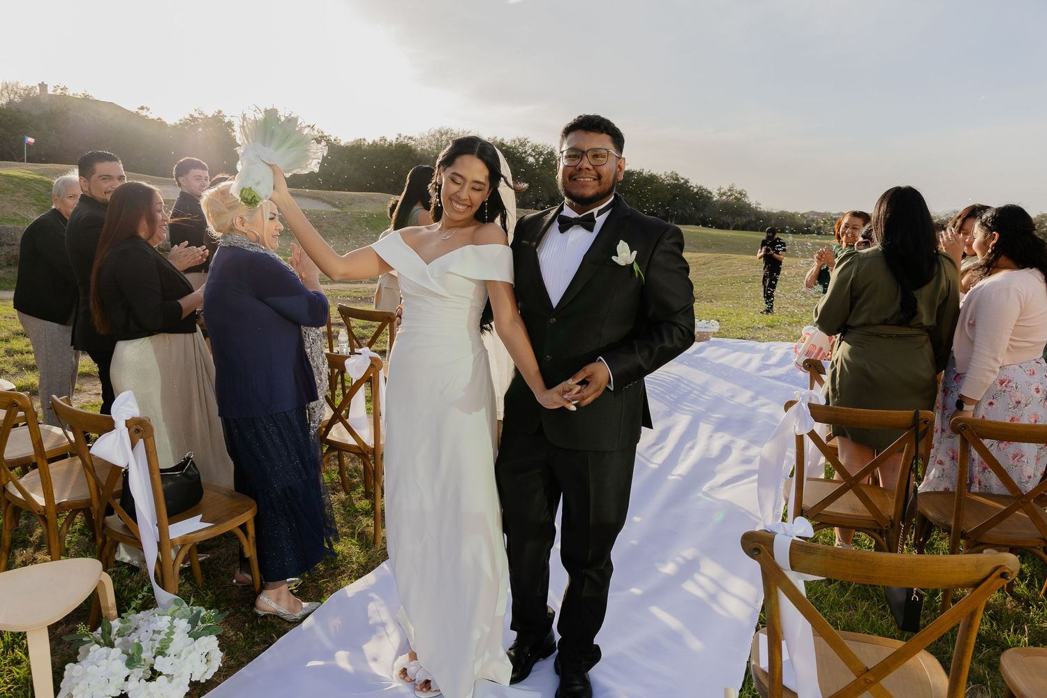 A newlywed couple walks back down the aisle at their outdoor wedding ceremony with guests seated on wooden chairs.
