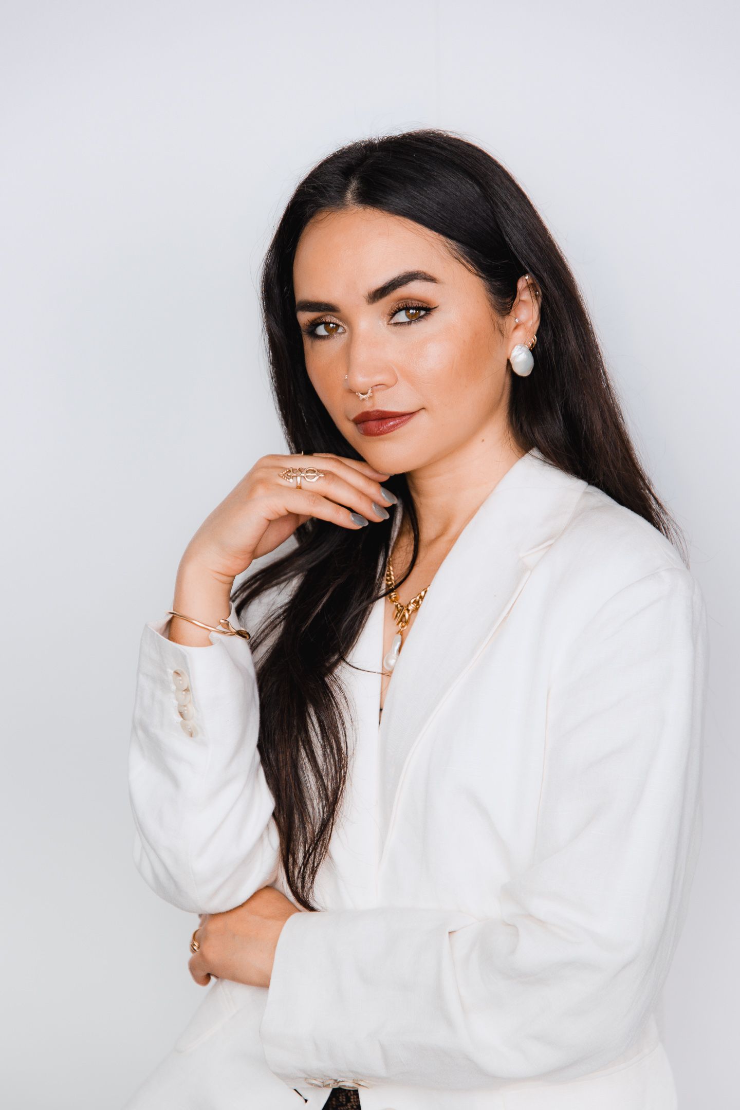A young woman with long dark hair wearing a white blazer and gold jewelry poses confidently against a light background.